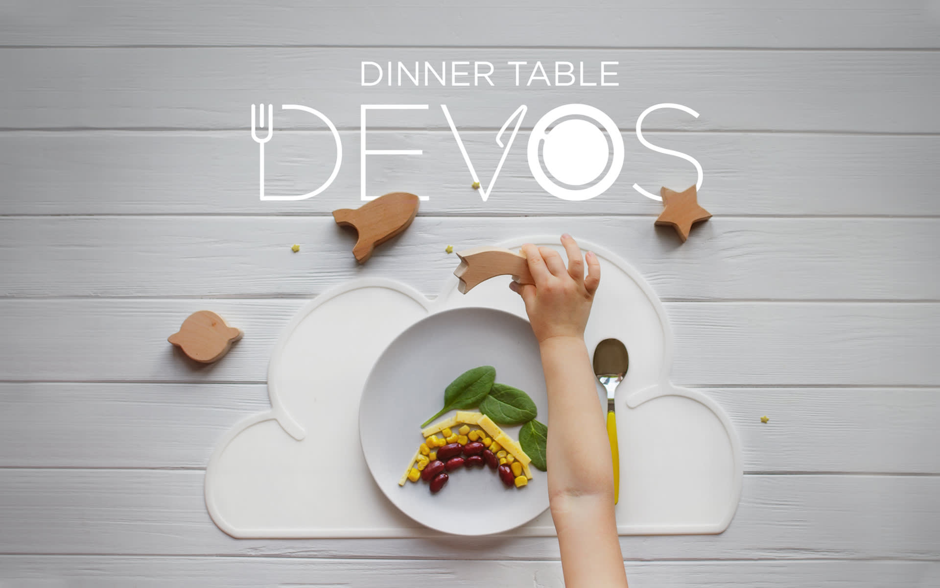 A young child reaches for a toy star near his dinner plate. In front of him is a white cloud shaped placemat, a white plate with several different coloured vegetables on it. Title page for Dinner Table Devos.