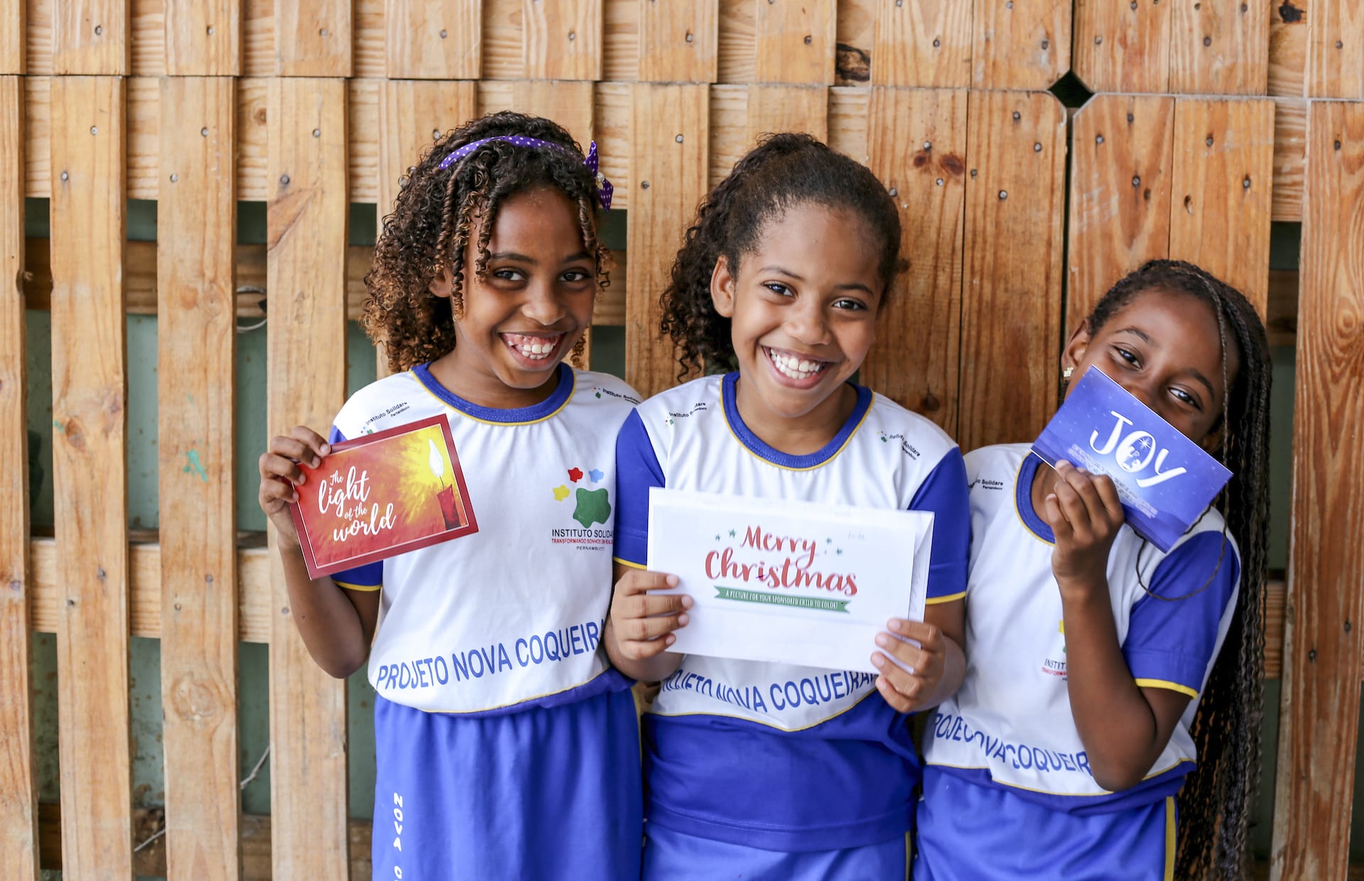 Three girls stand against a wooden fence holding up Christmas cards.