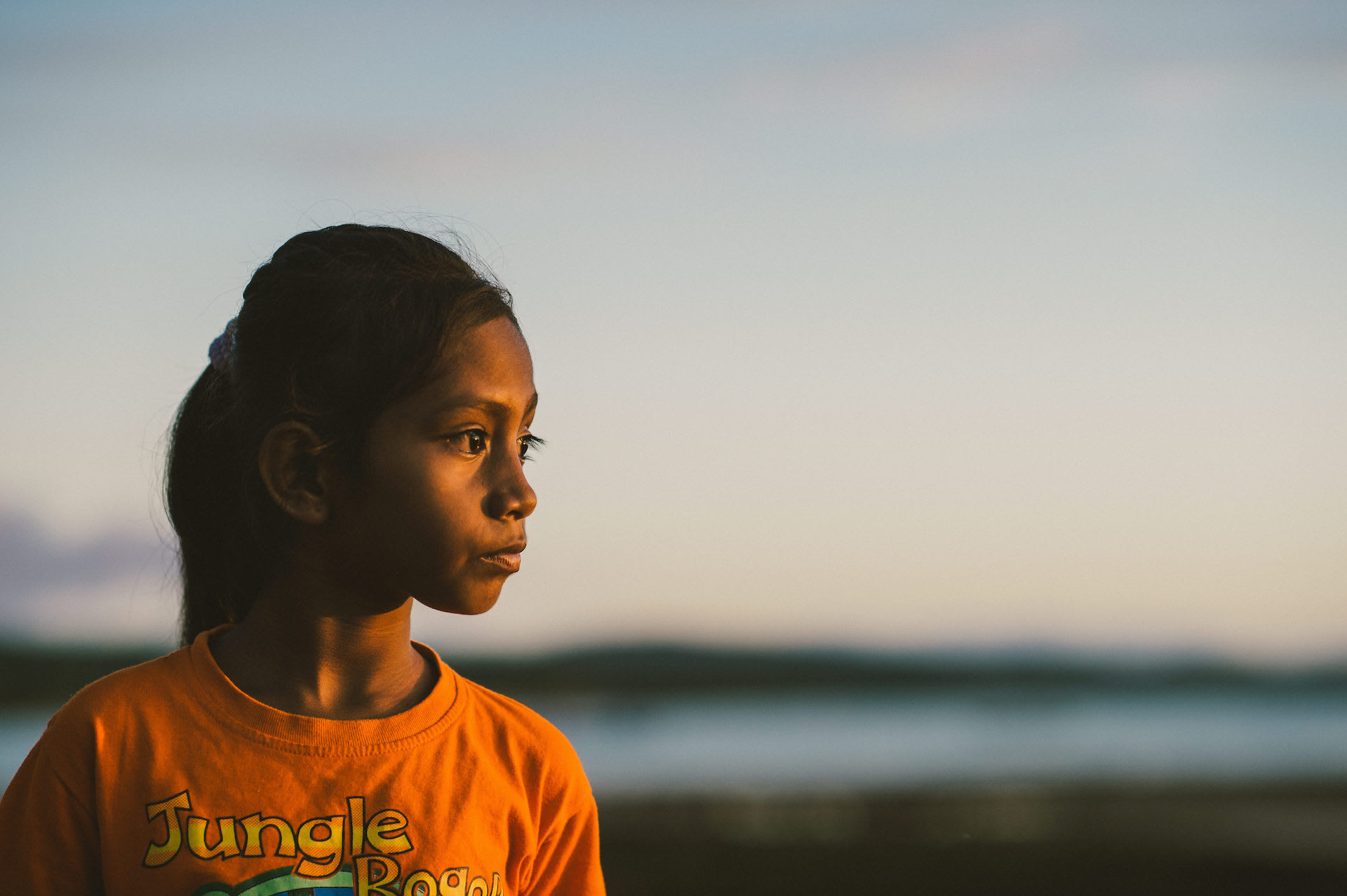 An Indonesian girl wearing an orange t-shirt and a pony tail looks at the sunset.