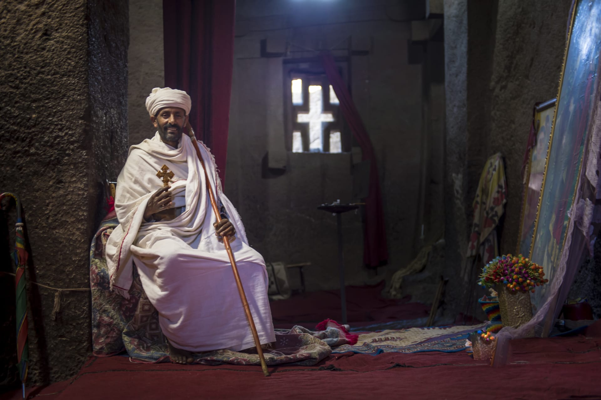 Ethiopian Orthodox Priest, adult male wearing a white robe sits by a collection of religious icons inside The Church of Saint George, one of many monolithic rock-cut churches hewn into the rocky hills of Lalibela, Ethiopia.