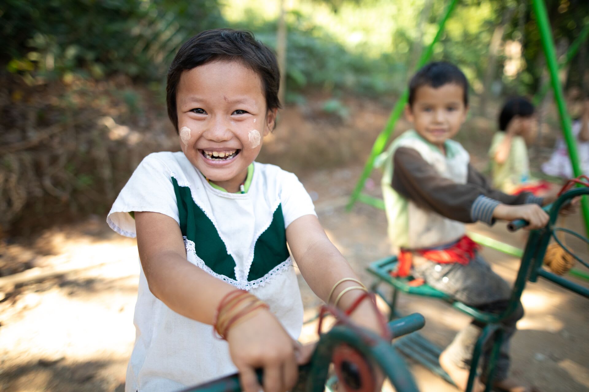 A Thai child in a white and green shirt plays on a playground structure. Other children are in the background.