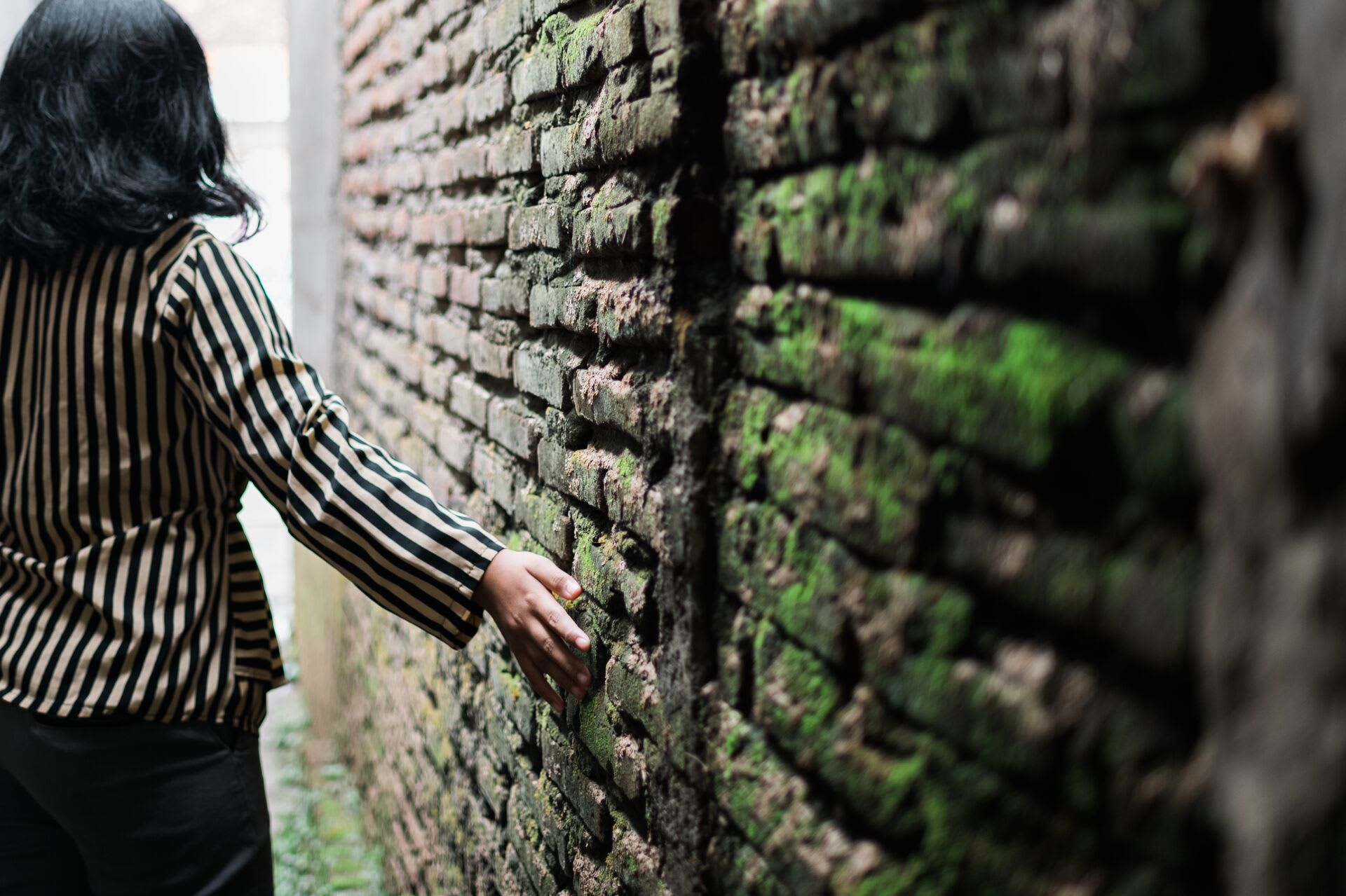 Girl walks down an alleyway with her hand running against the moss covered wall