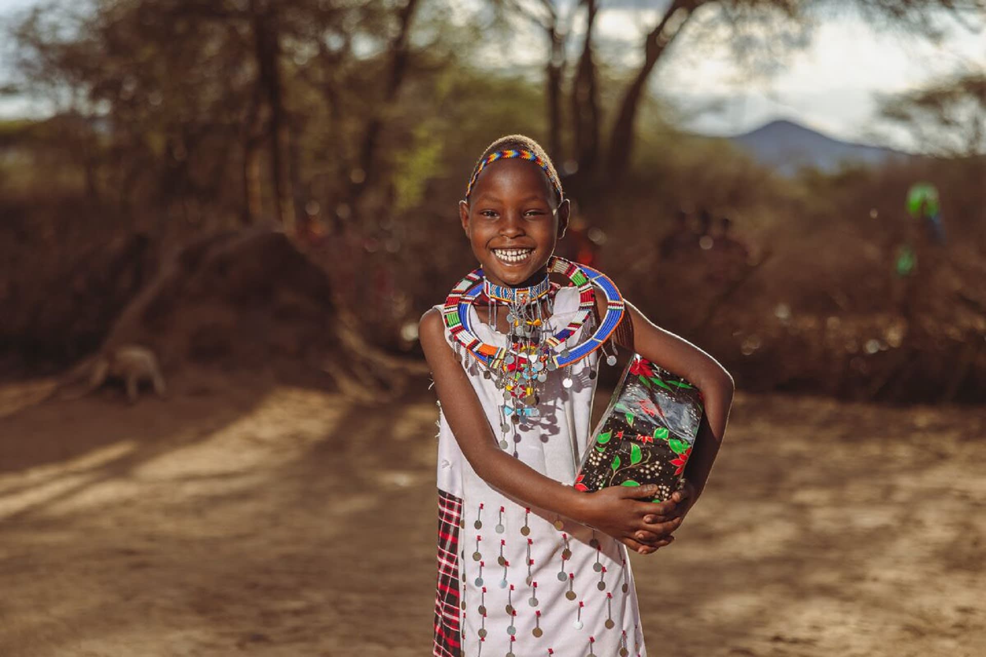 A girl is wearing red and white traditional clothing and is standing outside with trees in the background and holding a Christmas gift
