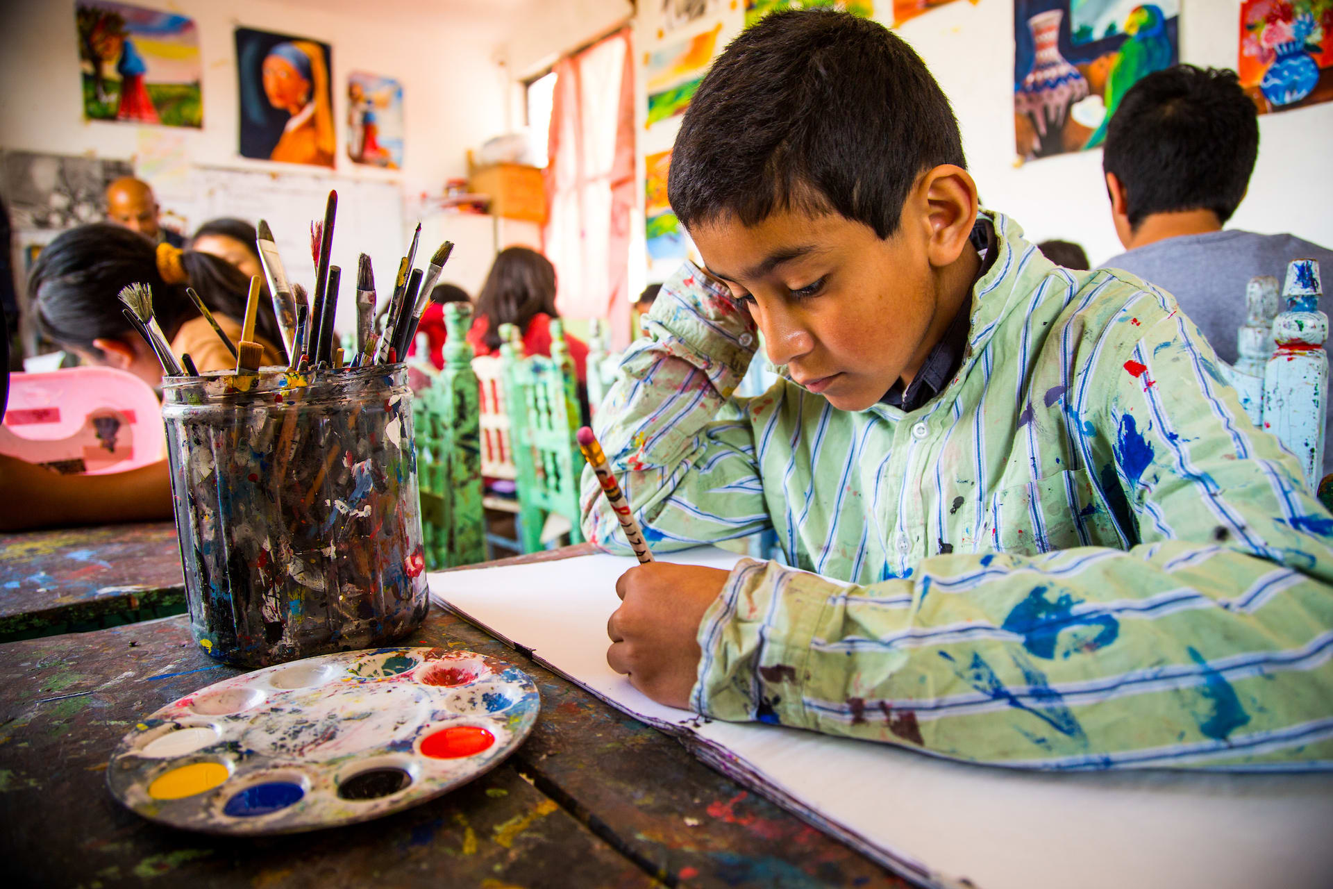 Hector, wearing a paint-smattered smock, sits at a table paining on a canvas.