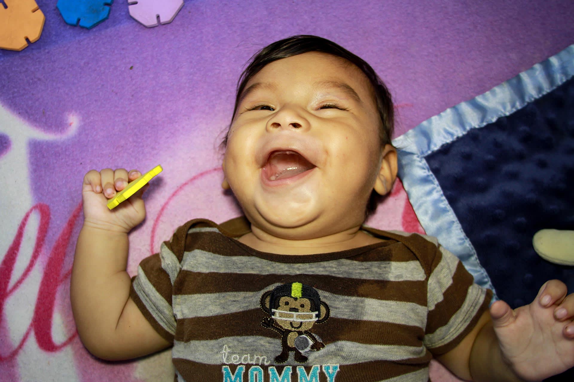A baby boy lies on a blanket and smiles a big smile.