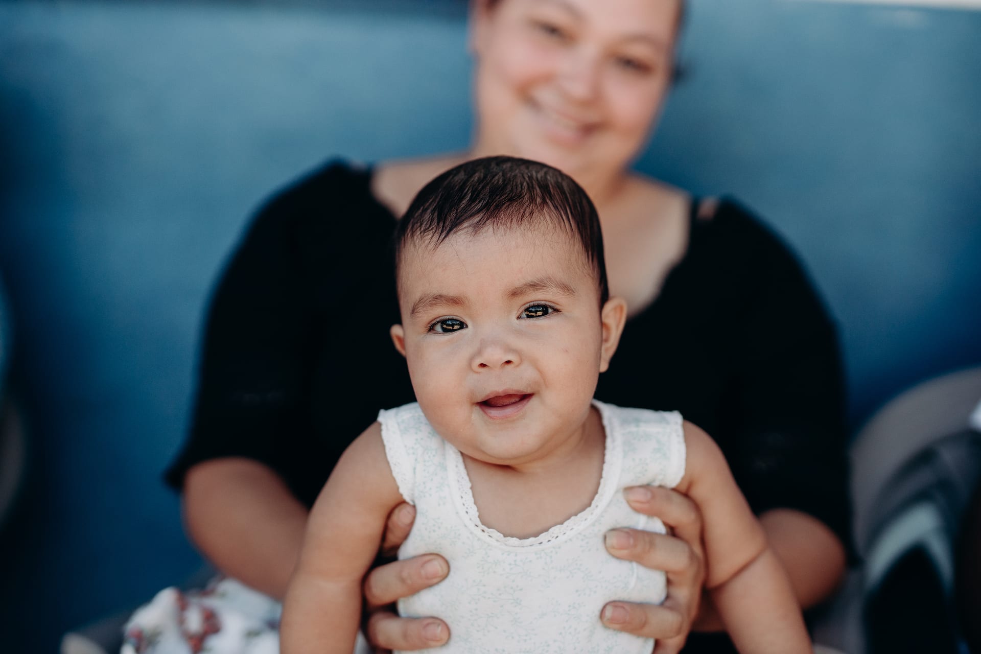Mother holds smiling baby girl up to the camera. She is wearing a white shirt. The background is blue.