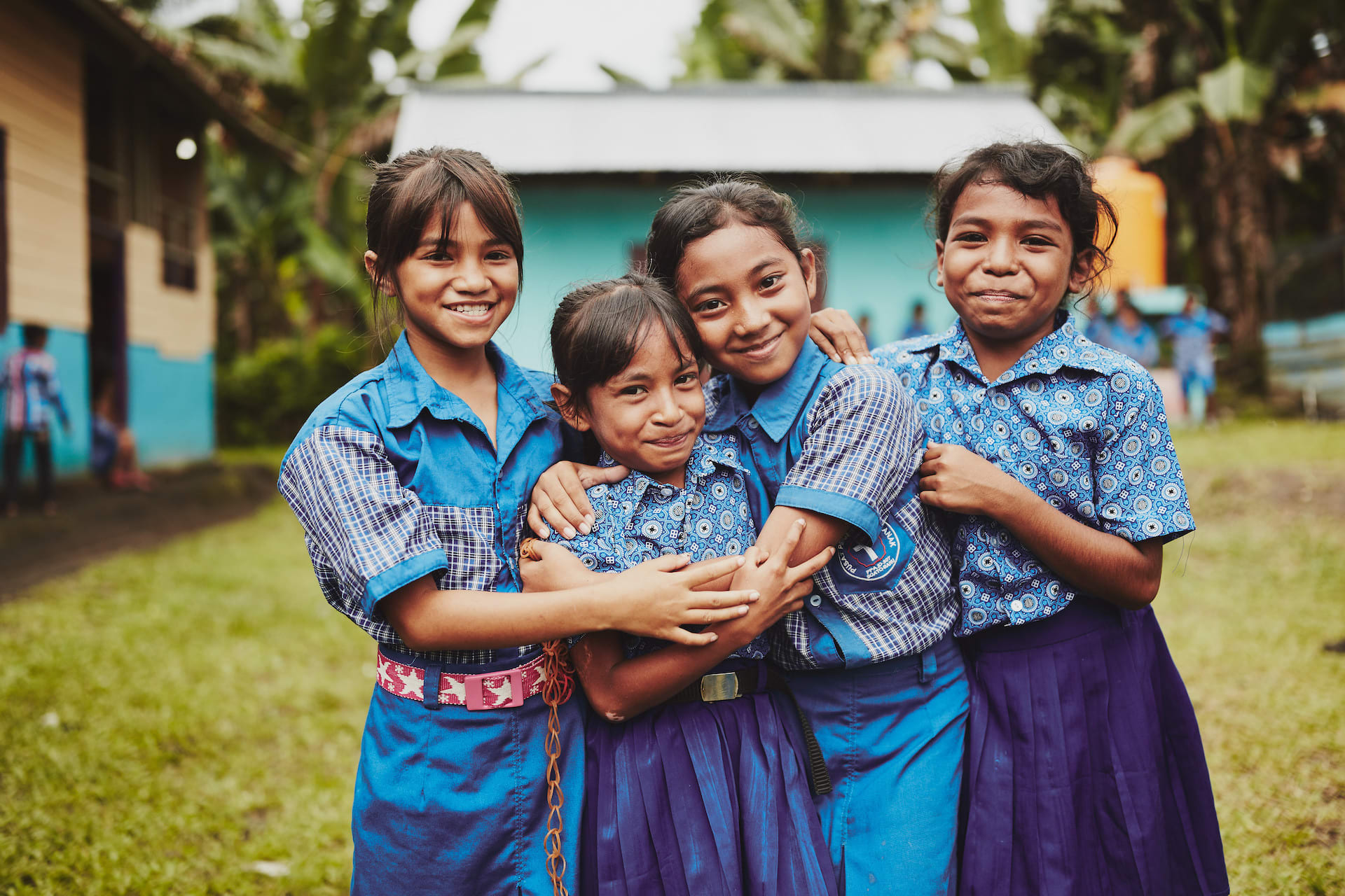 Four girls in blue school uniforms smile and hug one another.