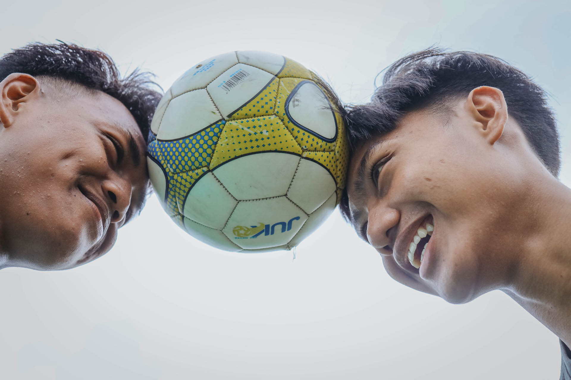 Two boys balance a soccer ball between their heads while smiling.