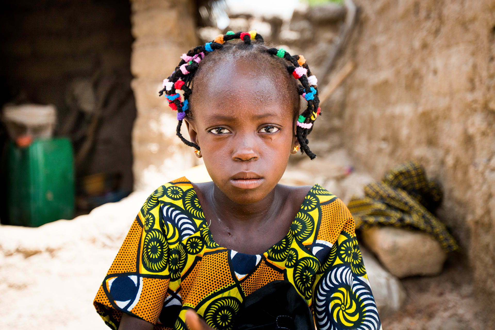 Young girl from Africa looks at the camera with a straight face. She is wearing a colourful dress and colourful beads in her hair.