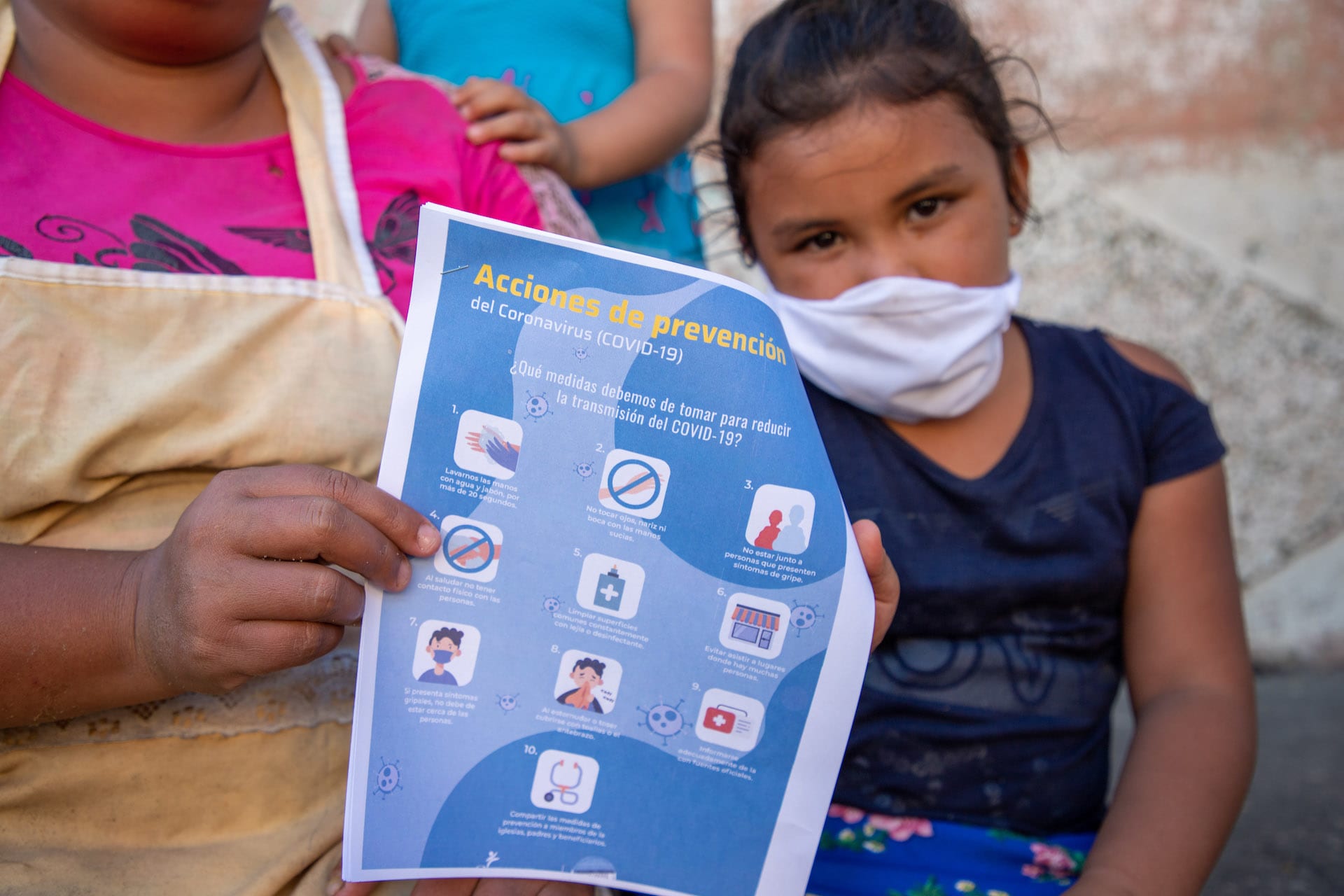 A girl wearing a mask sits next to her mother who is holding a pamphlet about COVID-19 prevention.