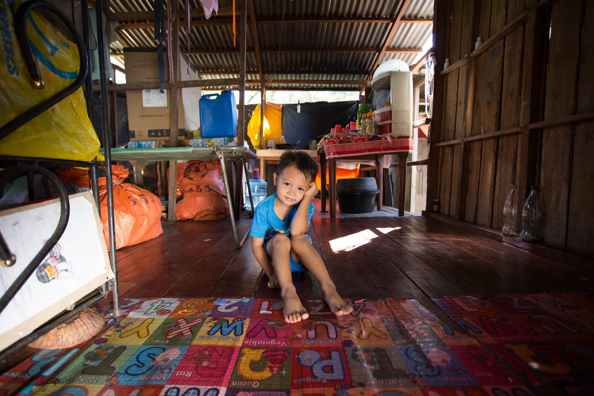 A young Filipino boy sits on the floor of his home. His legs are straight out in front of him and his head leans on his arm. He is wearing a blue t-shirt.