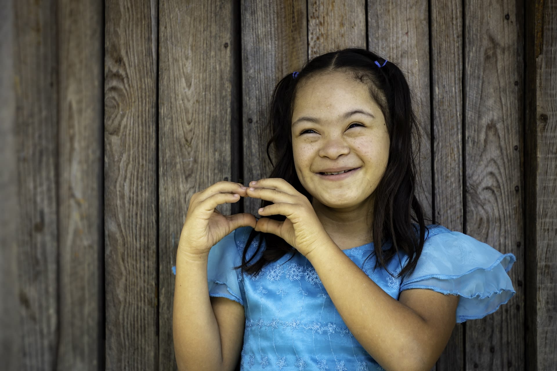 Little girl smiles wearing a blue dress and makes a heart with her hands. There is wood background behind her.