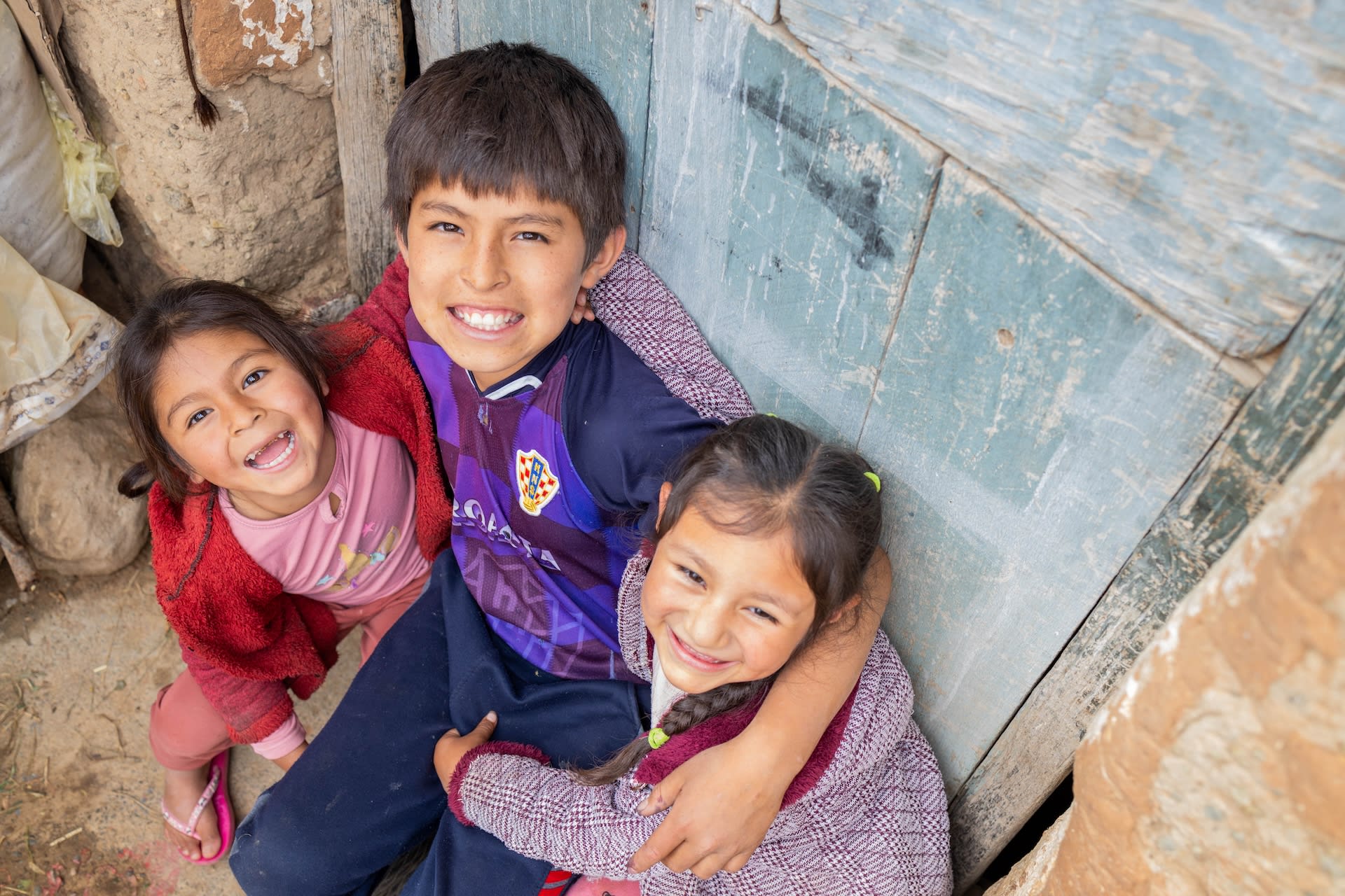 Three children looking up with big smiles. They are leaning back against a wooden wall.