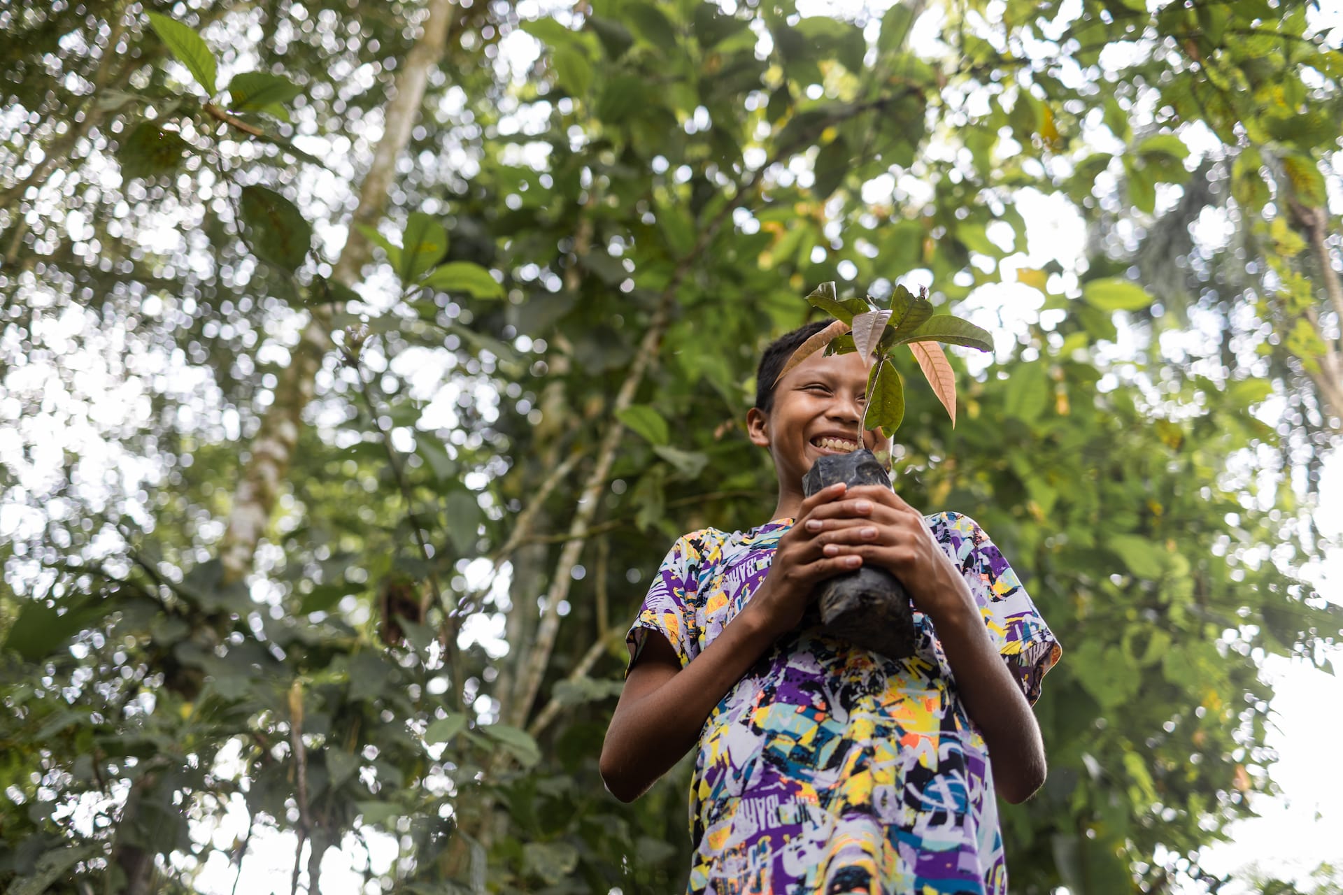 Waira, a 12-year-old boy in Ecuador, is standing in the jungle laughing and holding a plant.