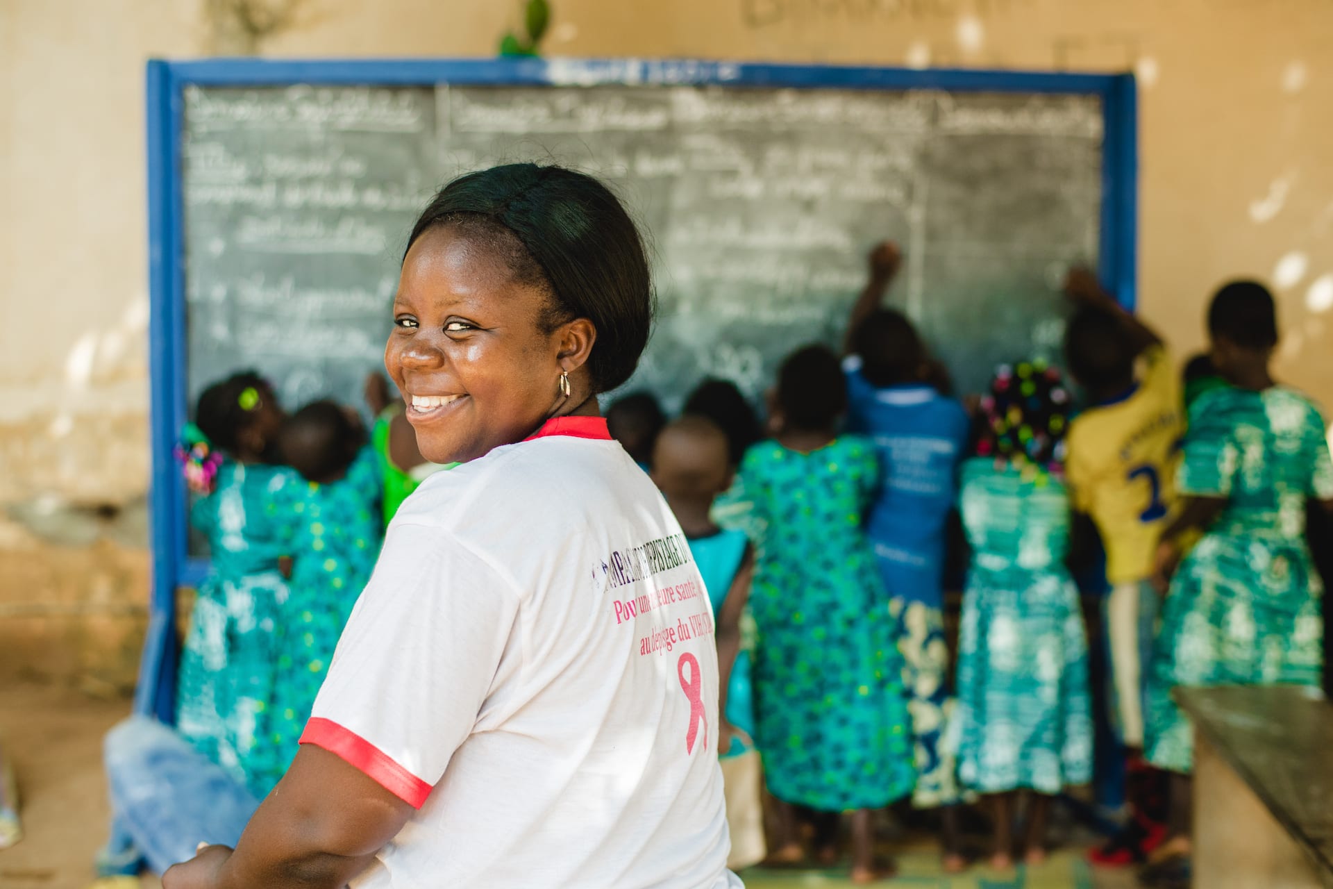 A woman in Togo sits in a classroom where children are working on a chalkboard in the background.