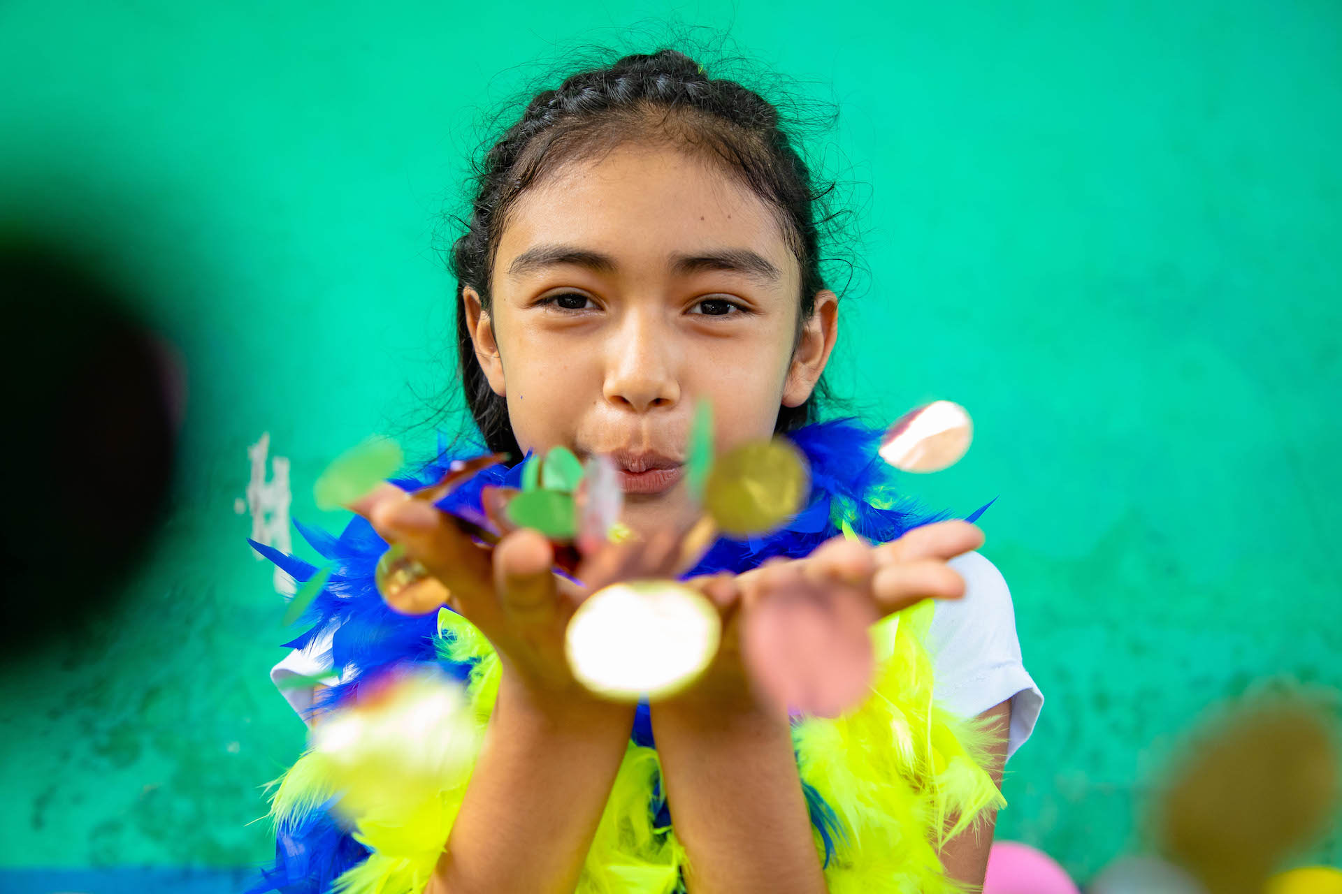 Genesis is wearing a white shirt and is blowing confetti toward the camera. She is standing in front of a green wall.