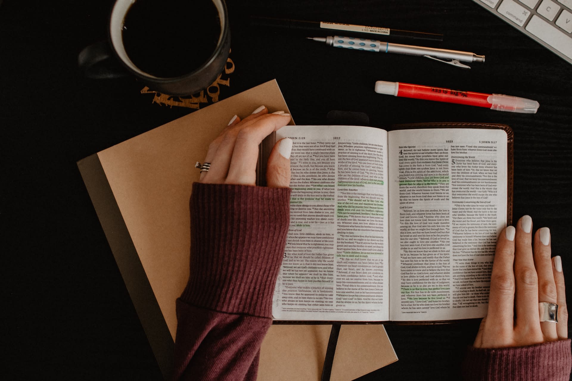 A Bible open on a desk, surrounded by a full coffee mug, pens and a journal. A pair of hands holds the Bible open.