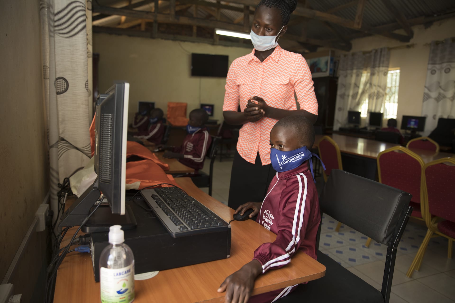 A young student works at a computer with a teacher standing behind her. Both are wearing protective face masks.