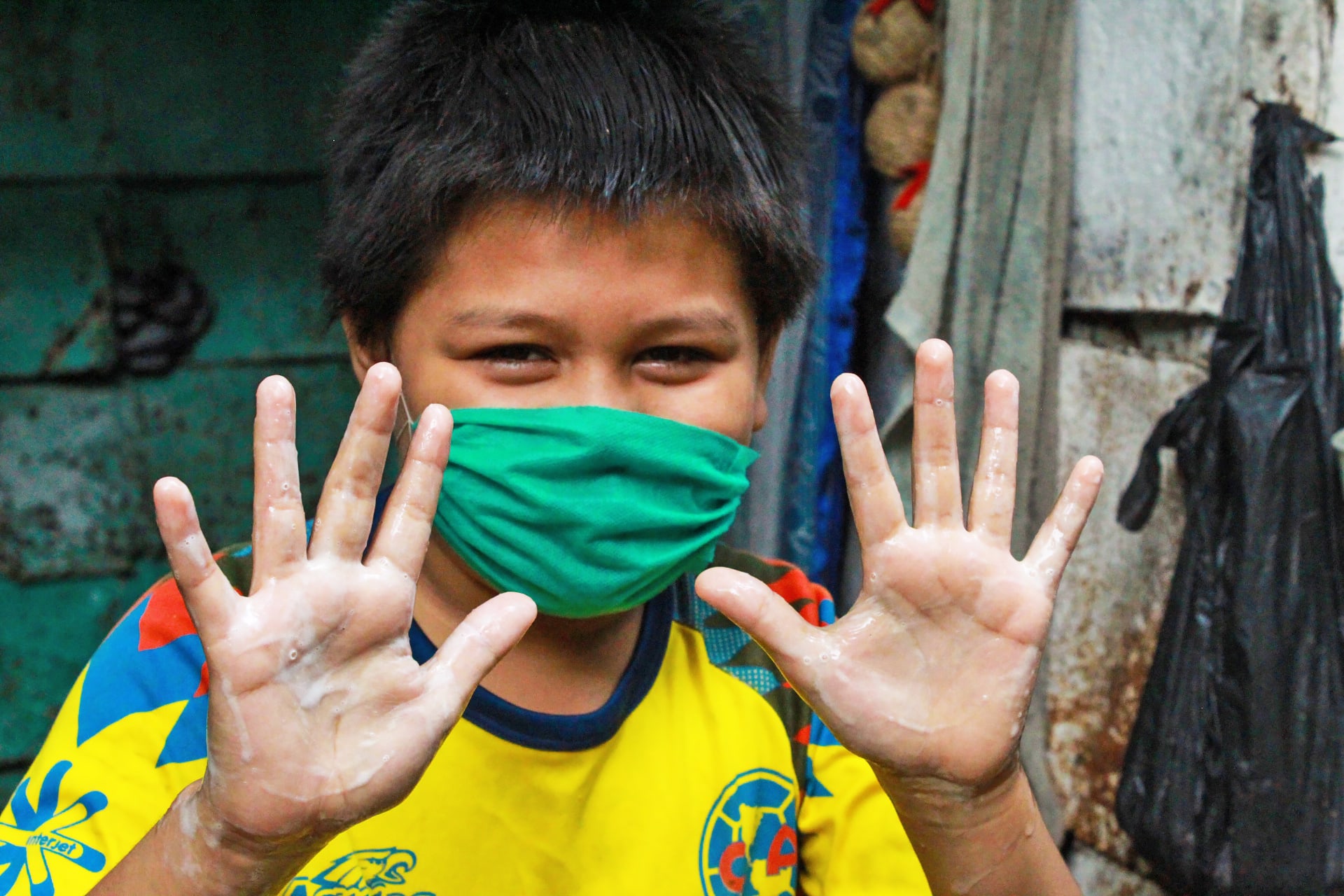 Ten-year-old Carmelo wears a green mask and yellow shirt and holds up both his hands to show they are freshly washed.