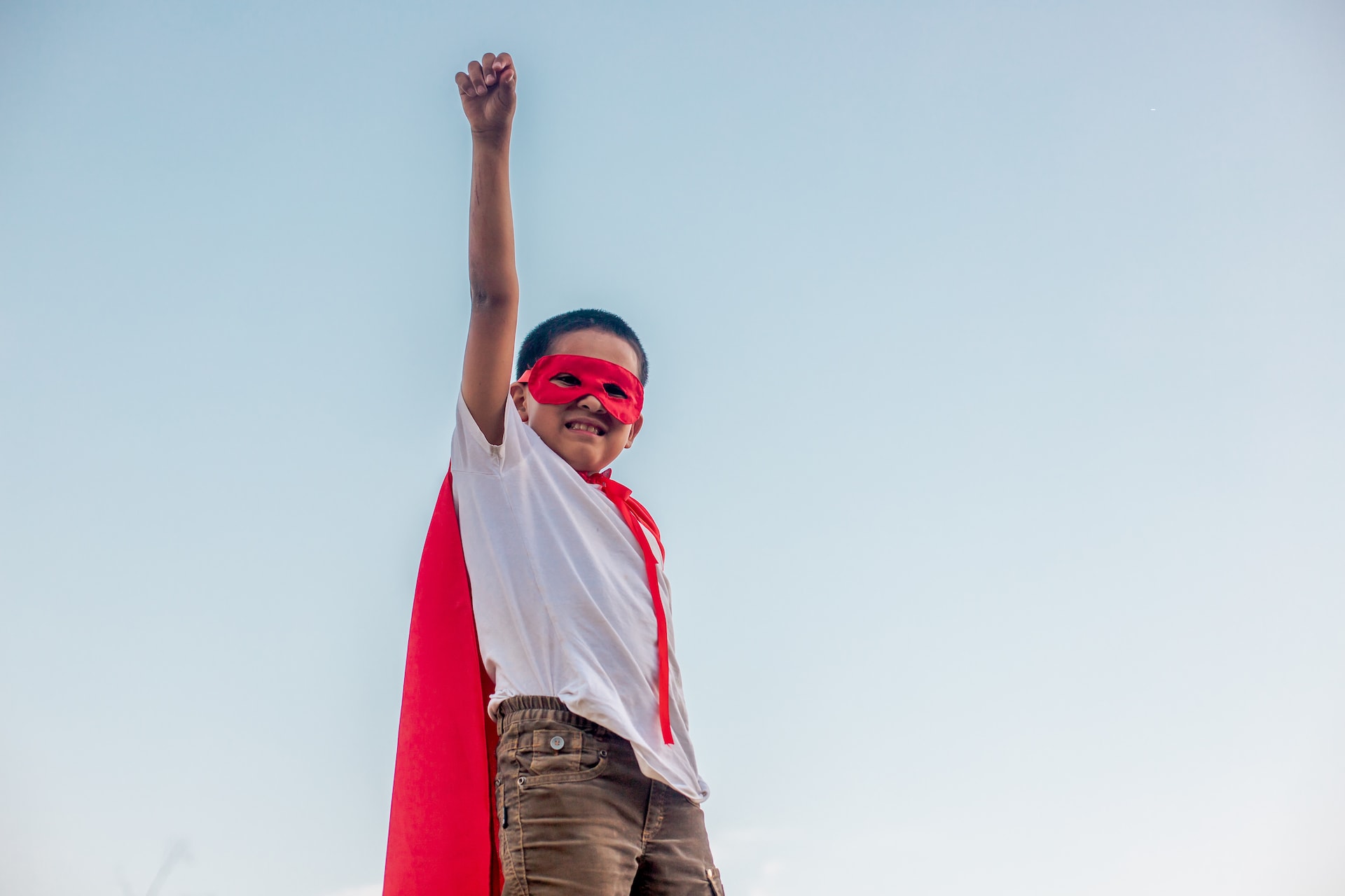Samuel, an 8-year-old boy from Bolivia, is wearing a white shirt, gray jeans, and a red cape. He is standing outside with one arm up in the air.