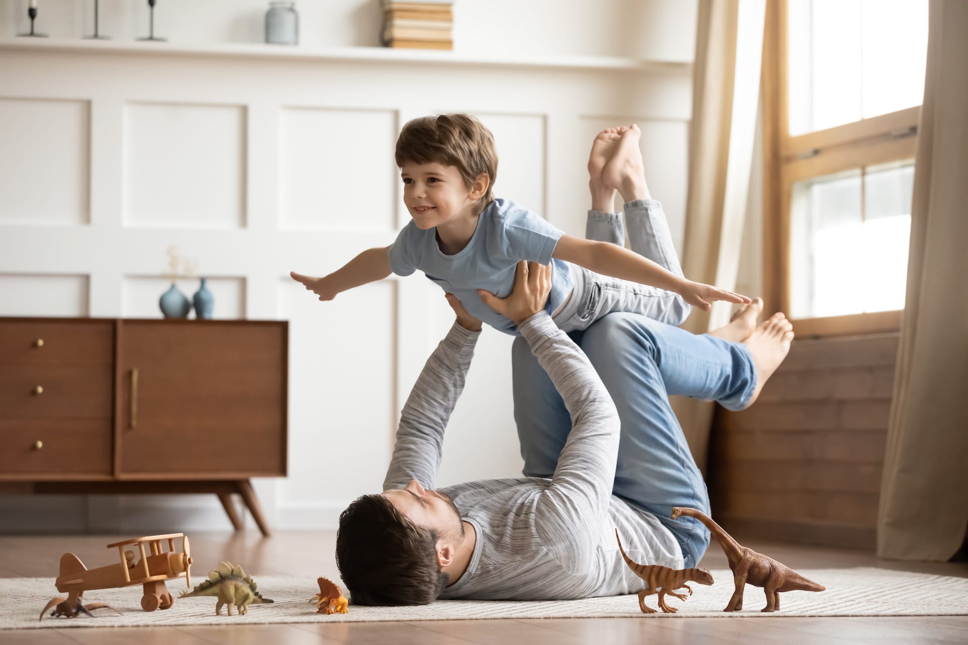 A father lying on the floor of a living room lifting up his young son as they play together.