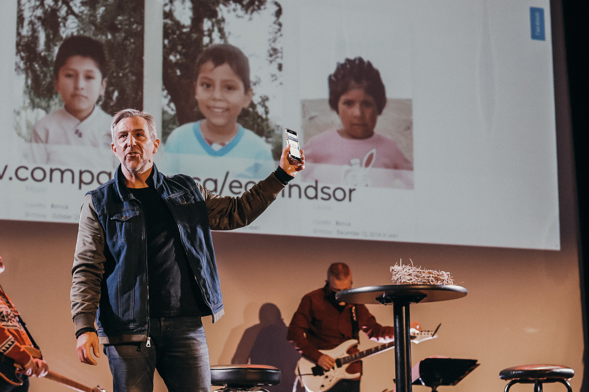 Pastor Brad stands on stage during a church service holding up a smart phone and inviting church members to sponsor a child with Compassion.
