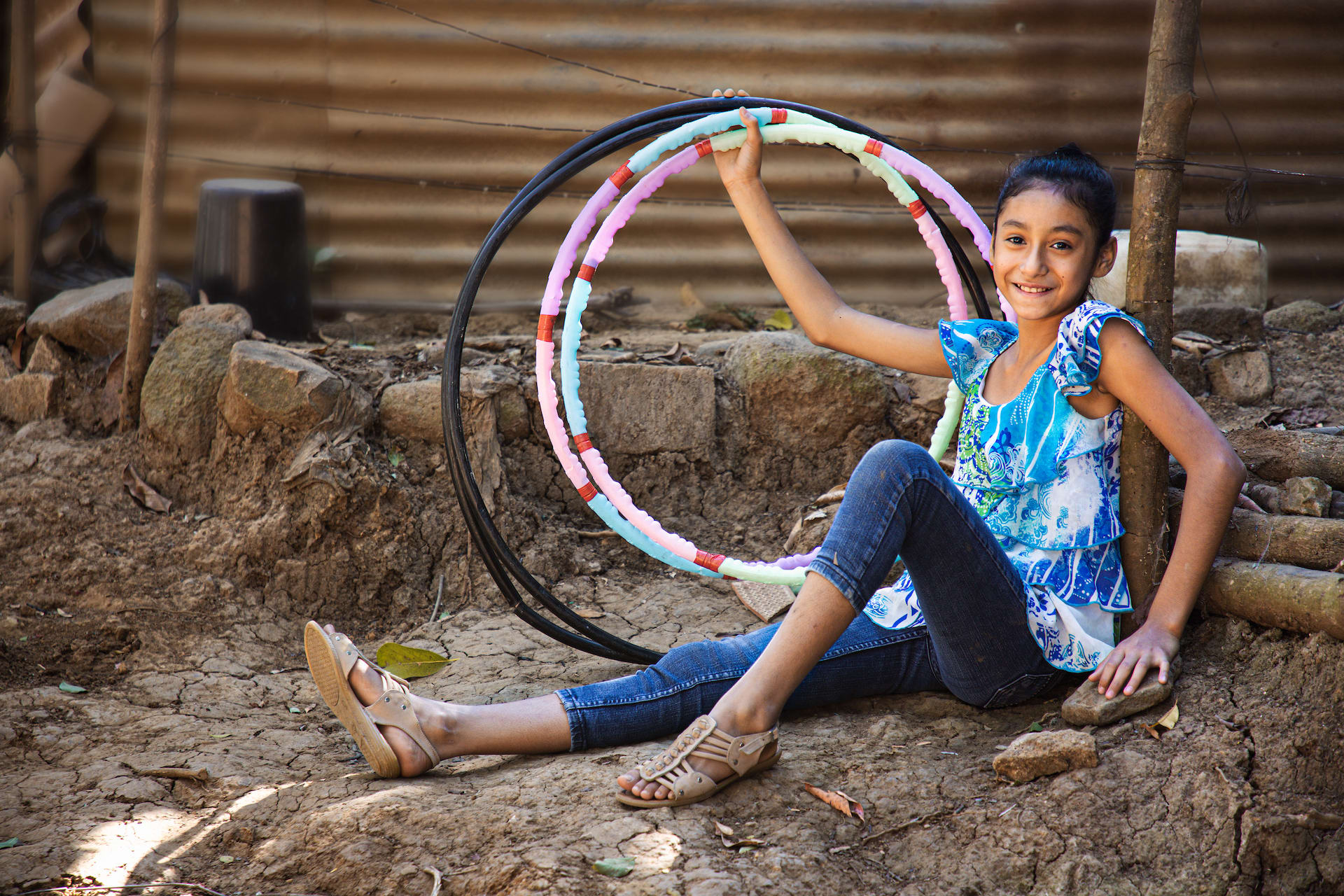 Suleyma is wearing jeans with a colorfully patterned shirt. She is sitting in front of her home and is holding several hula hoops.