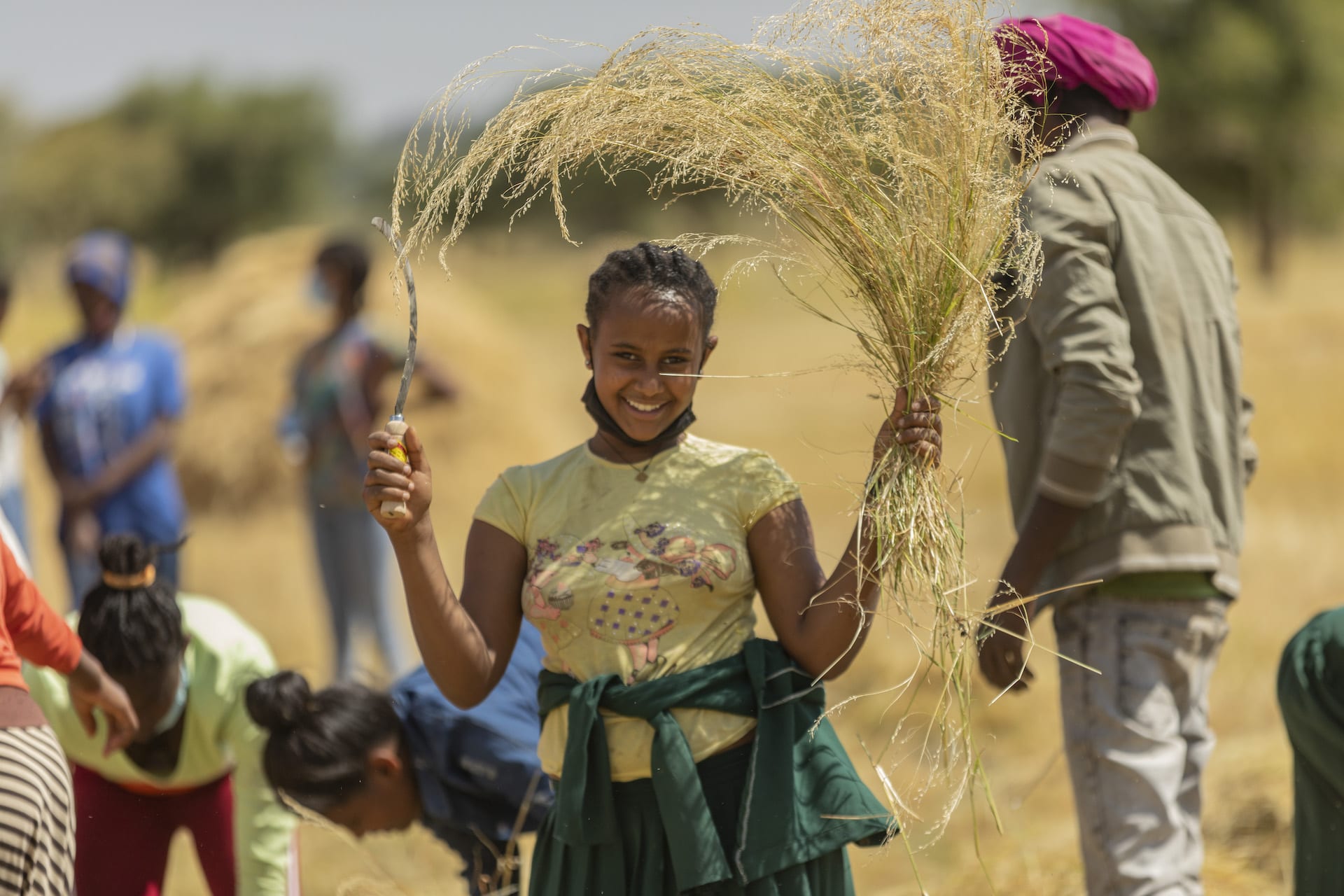 A teenage Ethiopian girl holds up a harvesting tool in one hand and a bunch of harvested wheat in the other. She is smiling and wearing a yellow t-shirt.