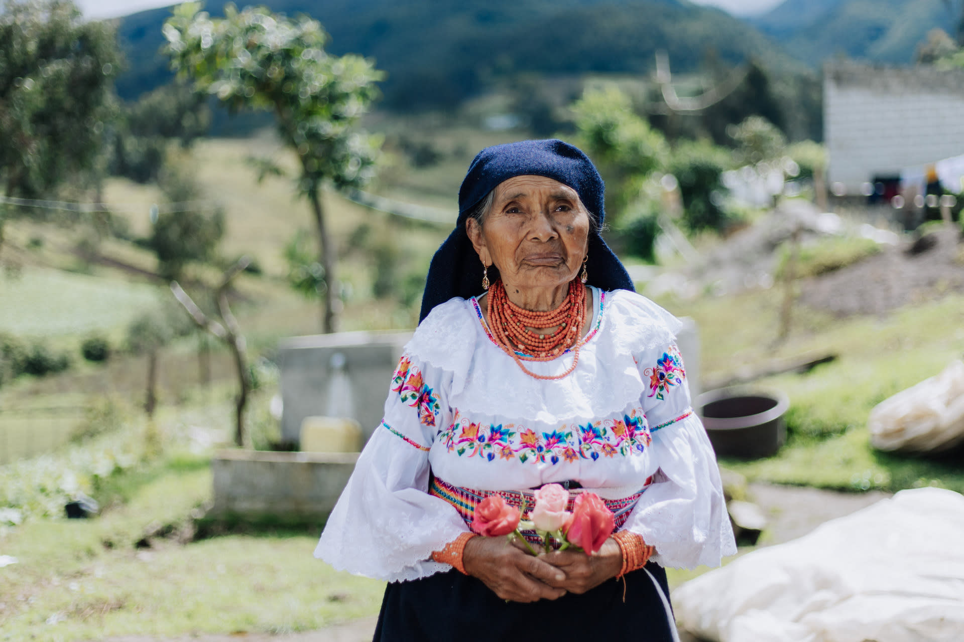 A 91-year-old Ecuadorian woman sits wearing traditional clothing and holding flowers in her hand. Behind her is a field of green grass and trees.
