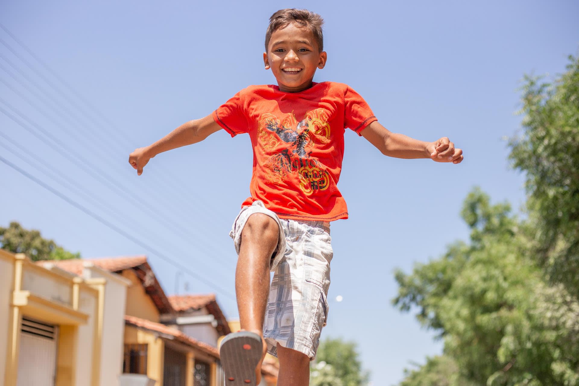 A boy earring an orange shirt and plaid shorts jumping.