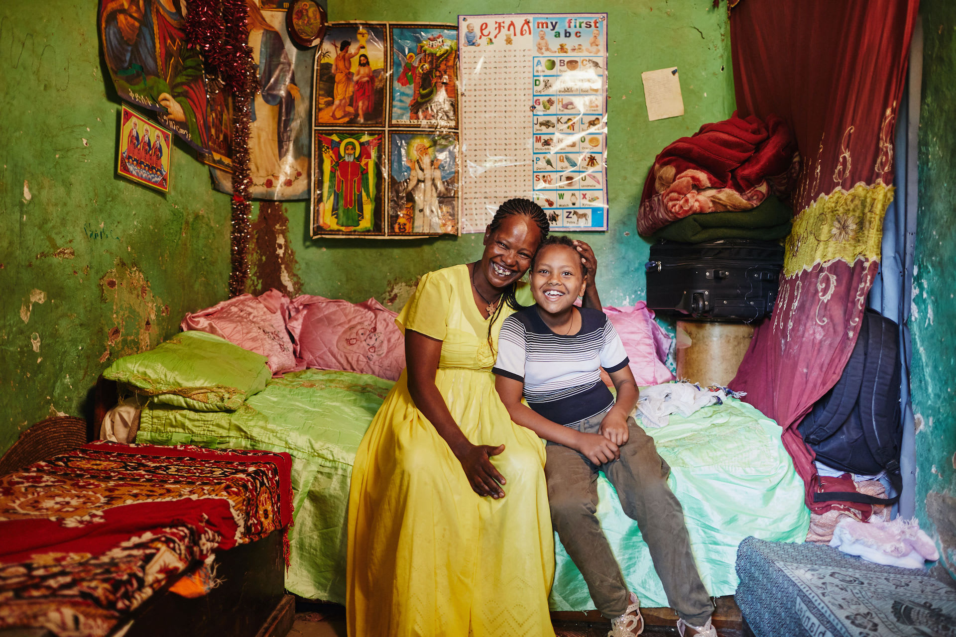 A mother and daughter sit on the bed and smile at the camera in their one-room home.
