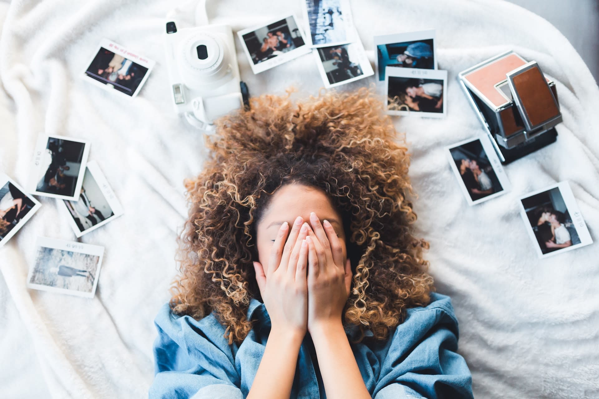 A woman lays on a bed with photos surrounding her head. Her hands are covering her face.