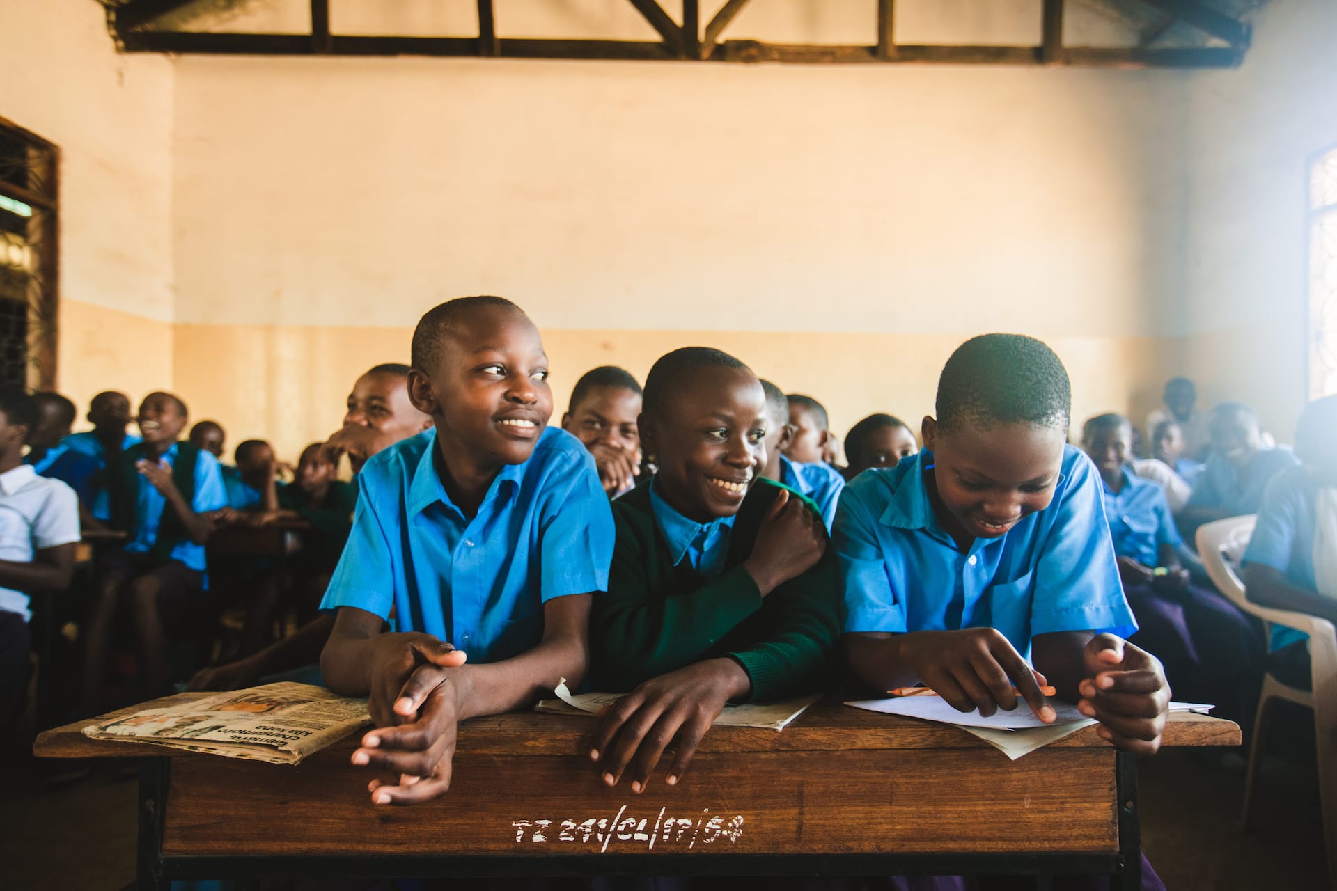 Three students sit at a long desk in a classroom. They are laughing together.