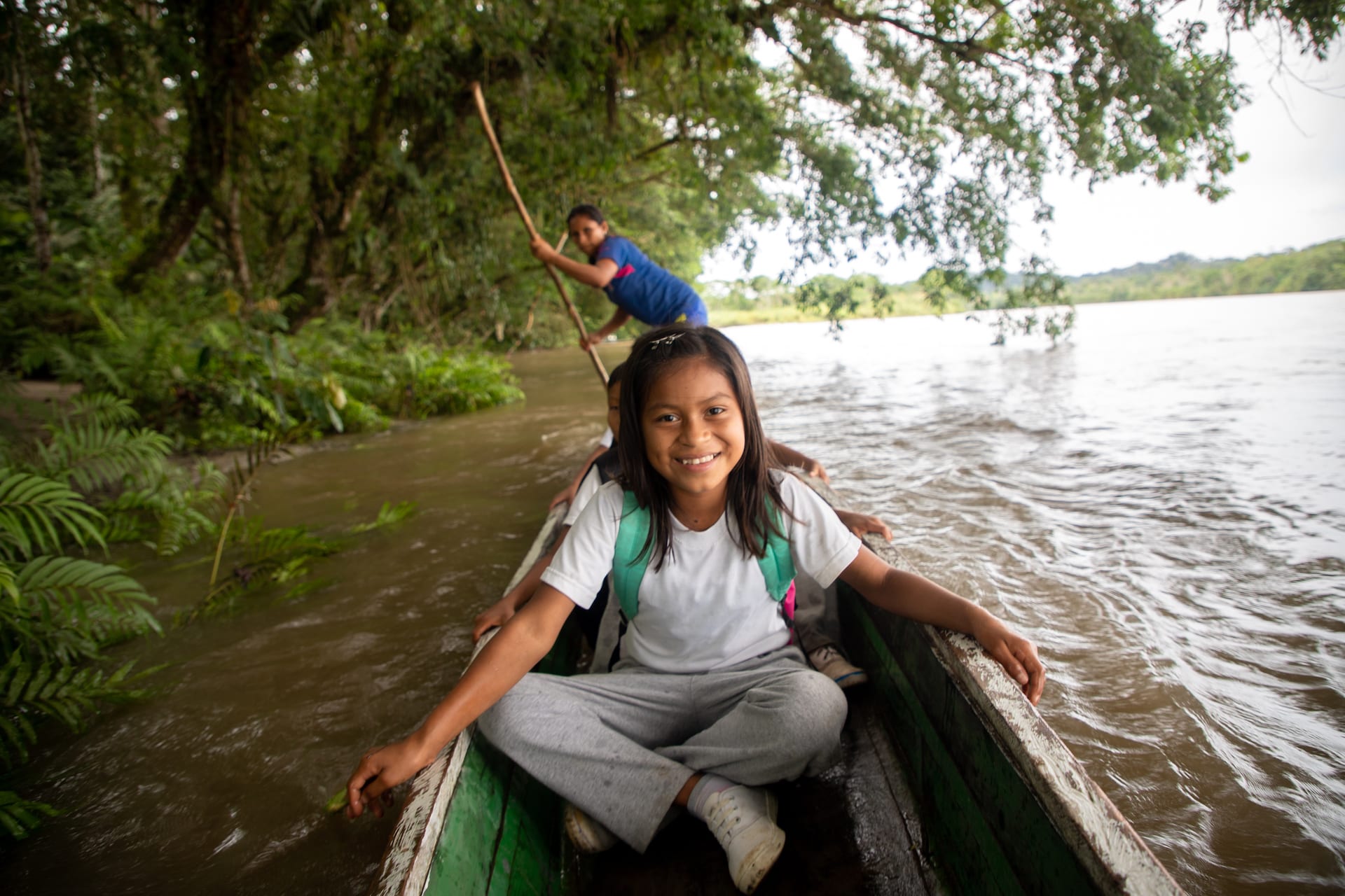 A young girl in a white tshirt sits in a canoe wearing a backpack, on her way to school.