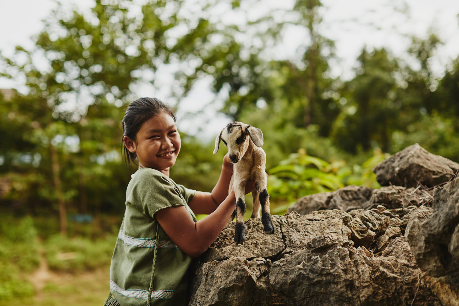 A young Thai girl in a green top holds a baby goat on top of a large boulder.