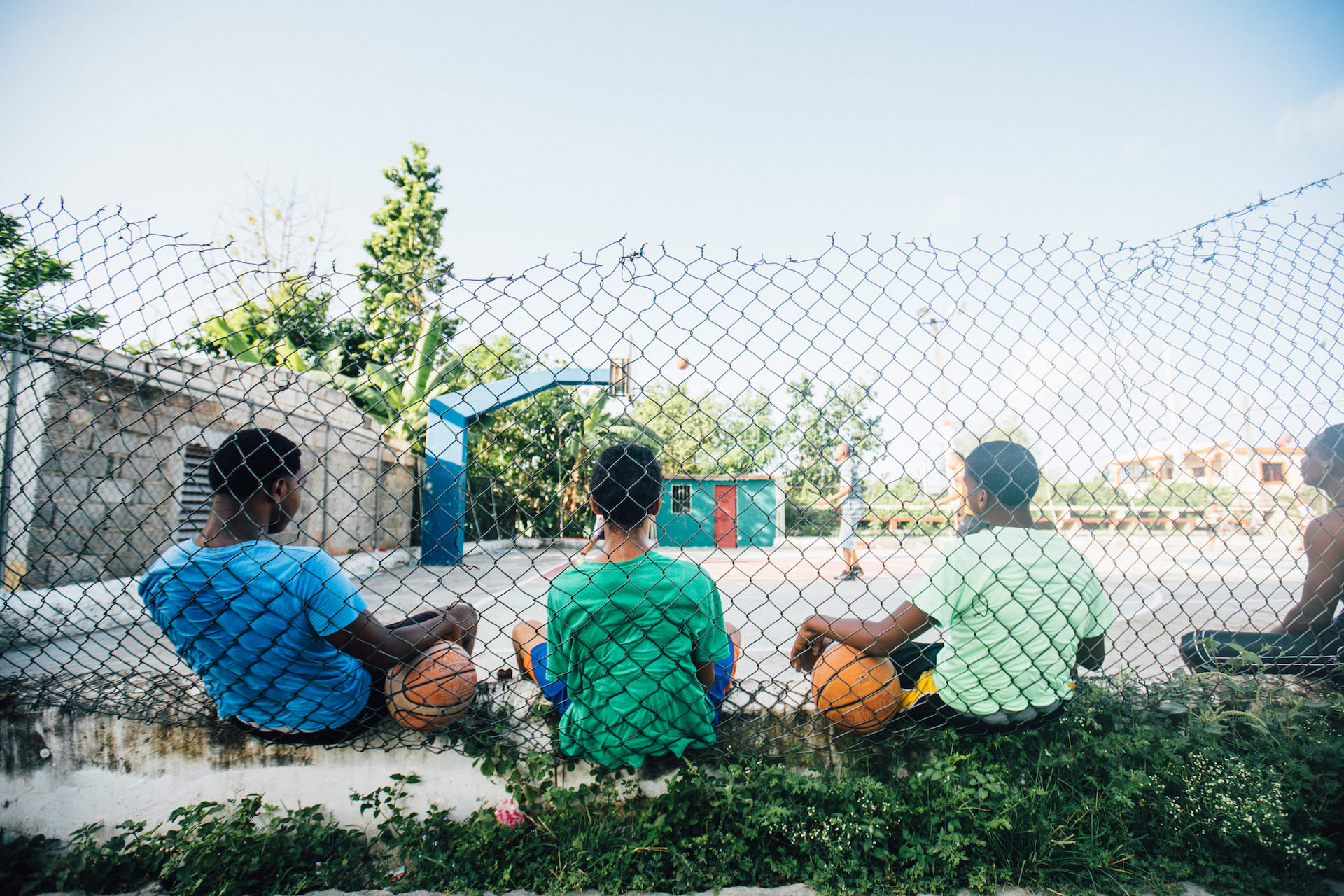 Three boys sit on the ground with their backs to the camera, leaning against a chain link fence. They are looking at a basketball court, holding basketballs.