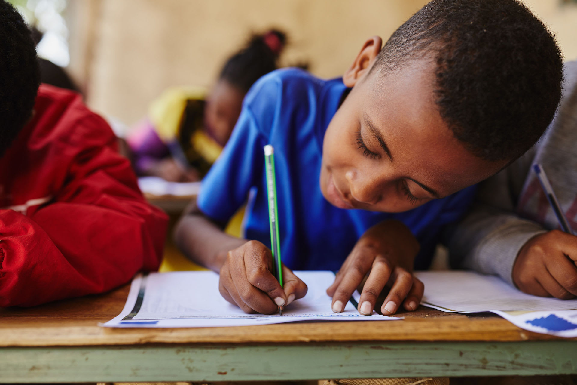 An Ethiopian boy in a blue shirt works on a letter to his sponsor. He is leaning over a wooden desk, writing with a green pencil.