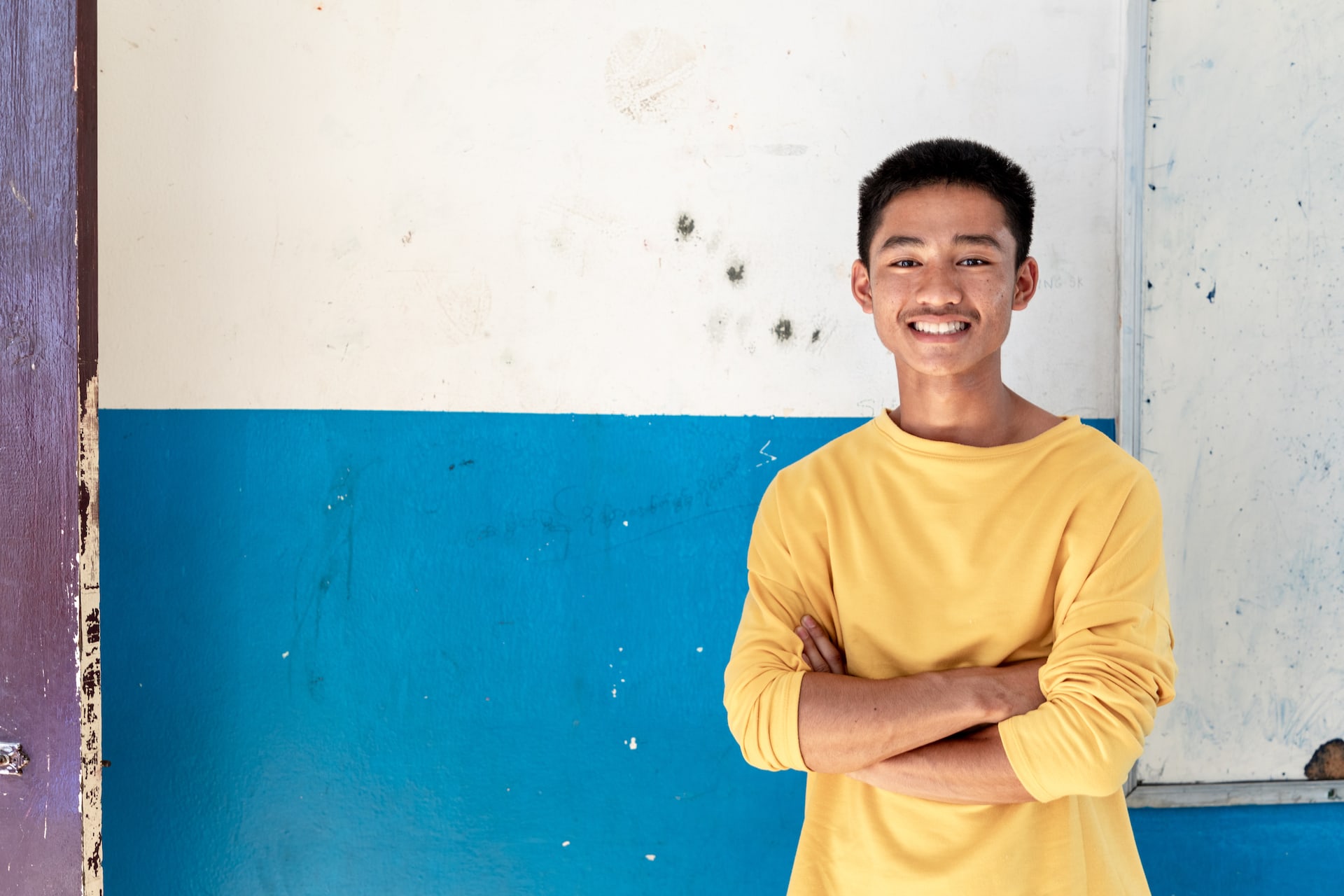 A portrait of 15-year-old Adun, a member of the soccer team from the Thai cave rescue. He is looking at the camera, smiling with his arms crossed. He is wearing a mustard yellow crew-neck long sleeve shirt.
