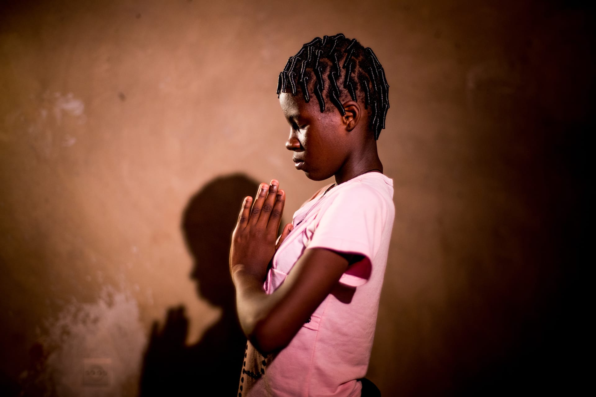 A girl in a pink shirt stands with her hands folded in front of her in prayer.