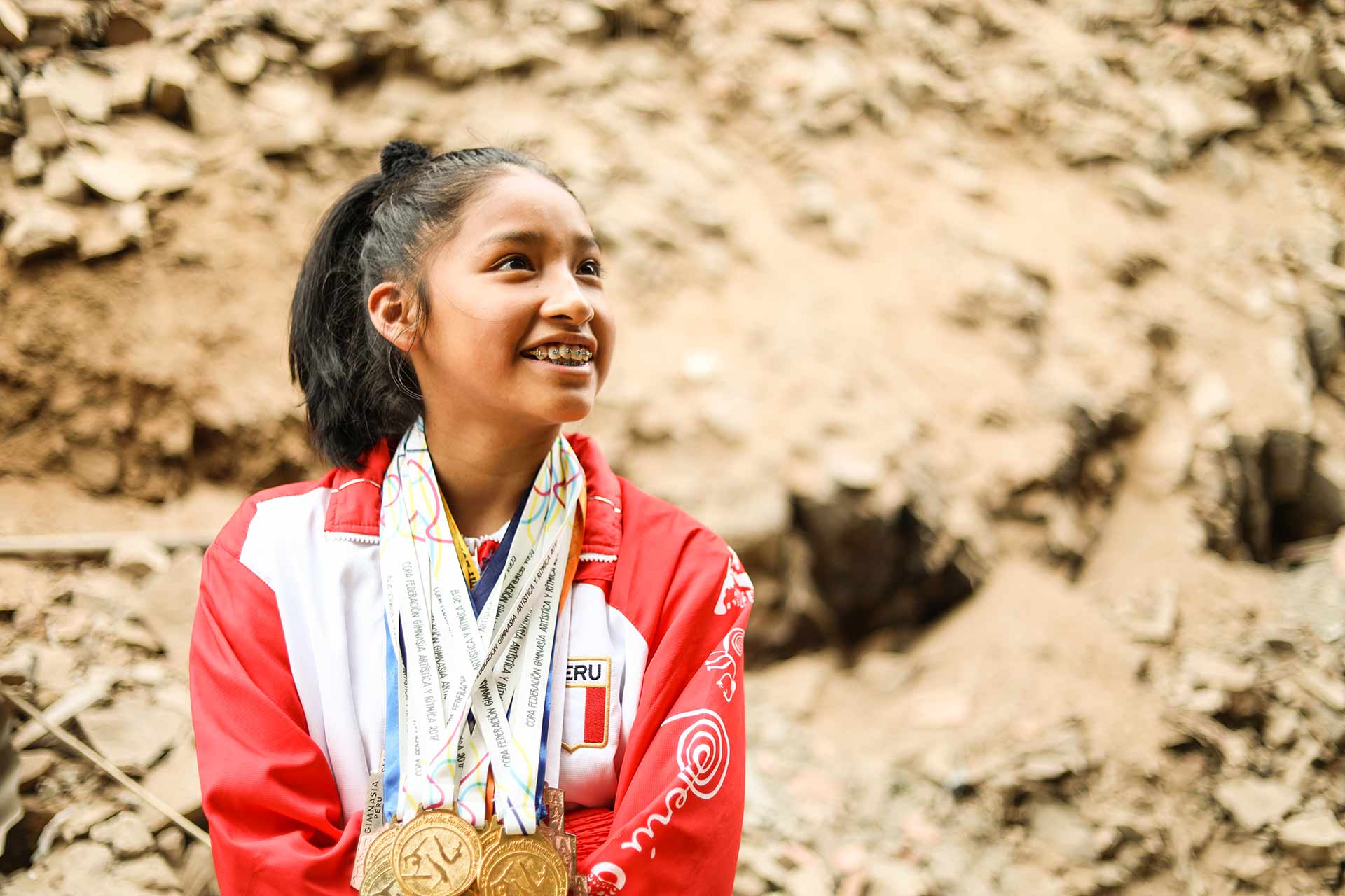 A girl wearing a red and white jacket has multiple gold medals around her neck. She smiles, looking up and to the side.