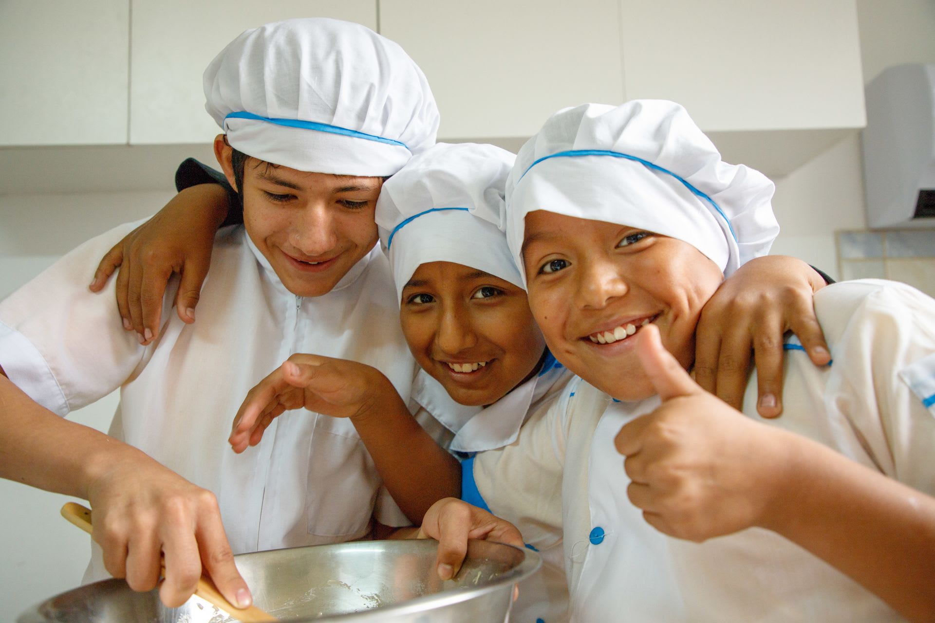 Three boys wearing baker's caps smile with their arms around each other, one giving a thumbs up while another holds a large kitchen bowl.