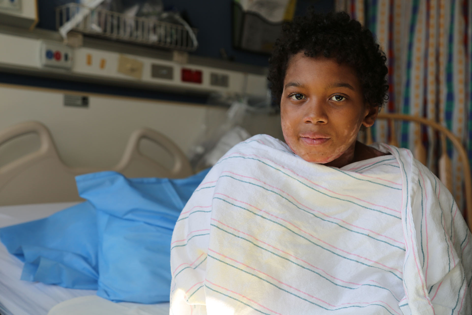 A boy sits in a hospital bed wrapped in a blanket