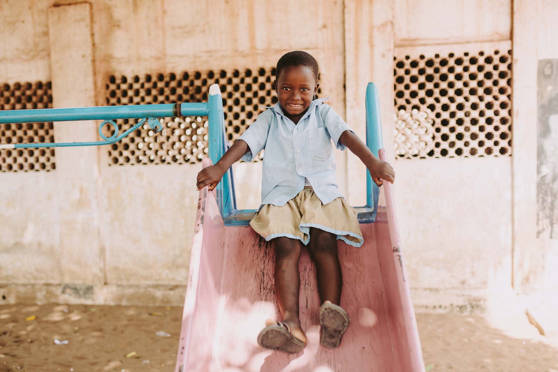 Aklobessi, girl, child, wearing a blue shirt, brown skirt, small earrings, smiles as she is playing, play time, play, sliding down the slide outside at the center on the playground equipment.
