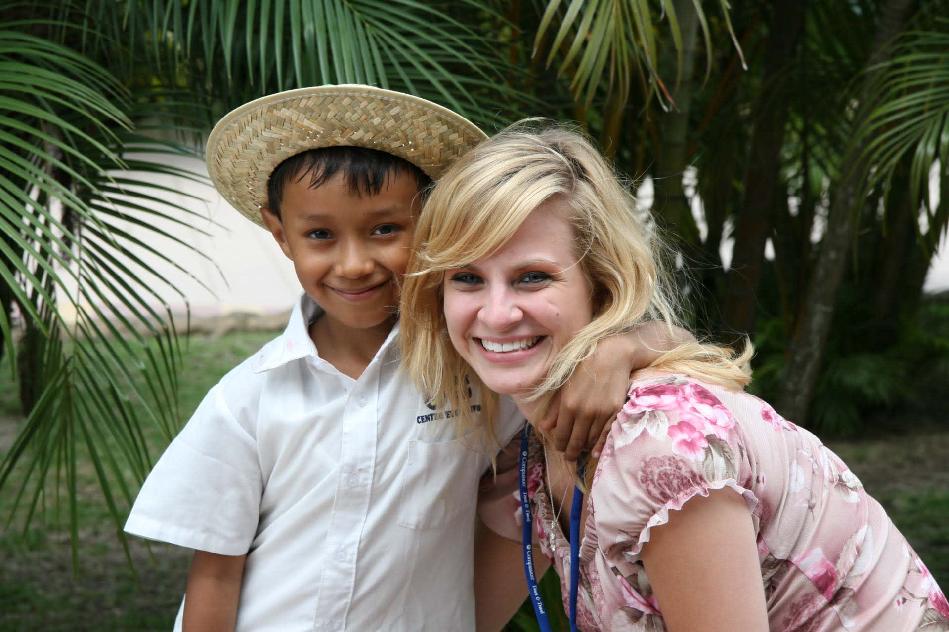 A boy and his sponsor are smiling together. They are standing together outside in front of the trees. The woman has blonde hair and the little boy is wearing a straw hat.