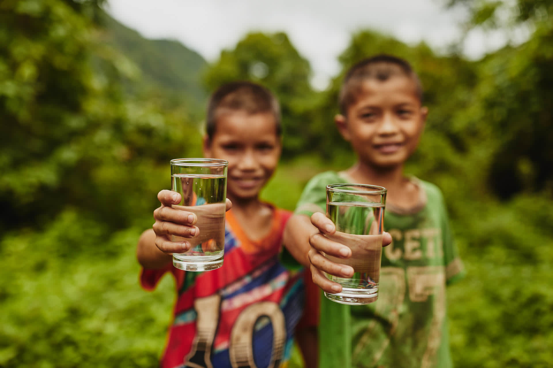 Two boys hold up clear glasses of water. The boys are out of focus and the background is lush jungle.