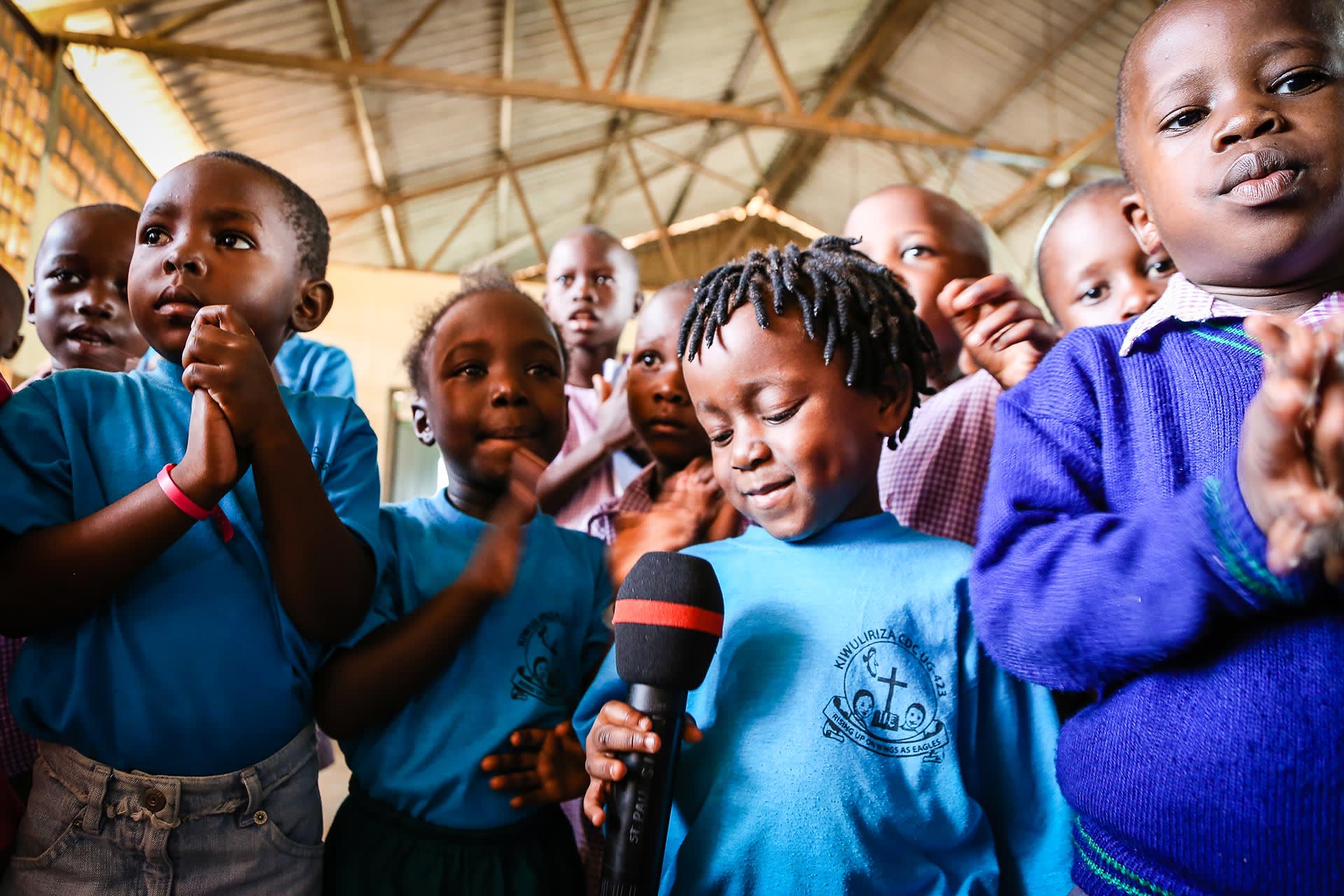 A child looks down at a microphone as other children crowd around.