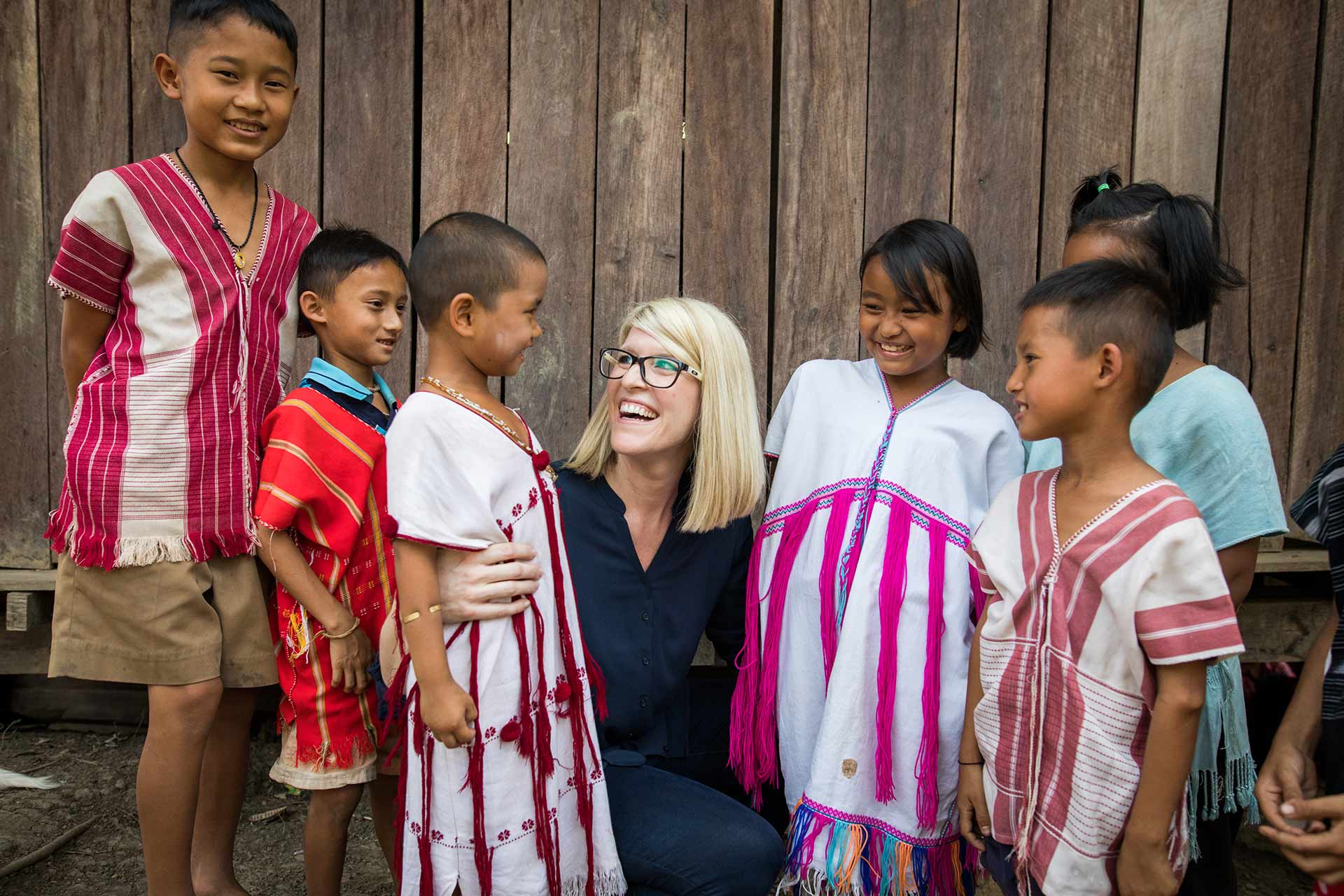 A blonde woman in a blue shirt crouches, surrounded by six children wearing colorful tribal shirts