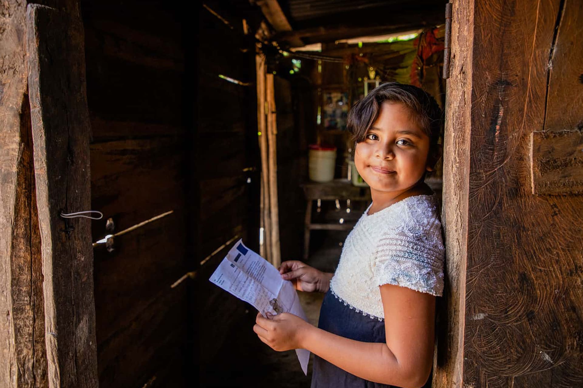 Little girl from Central America leans against a doorway smiling and holding a letter. She is wearing a blue and white shirt.