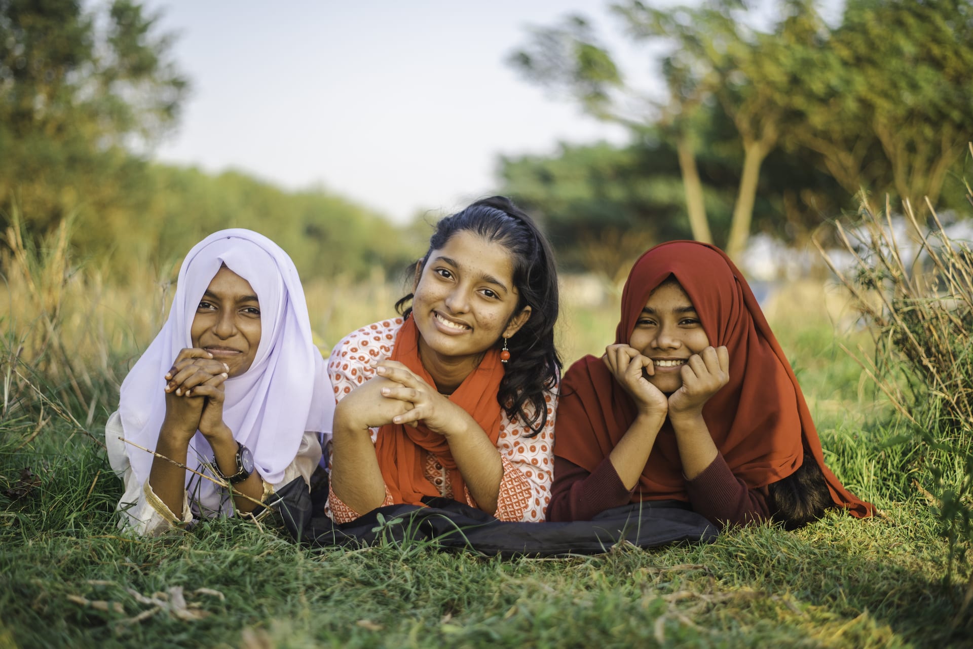 Jharna, Tisha, wearing a red and white outfit, and Munira, are laying down outside and there are trees in the background.
