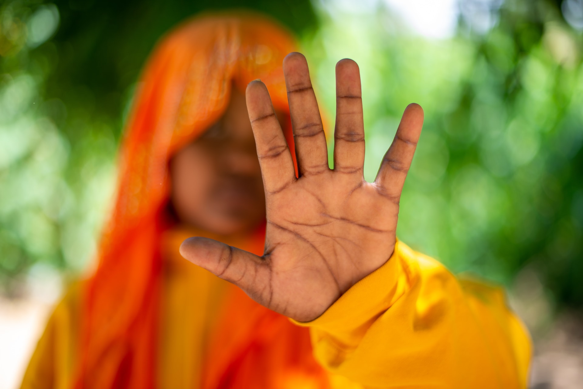 Sarah is wearing a yellow dress and an orange head covering. Her hand is stretched out toward the camera.